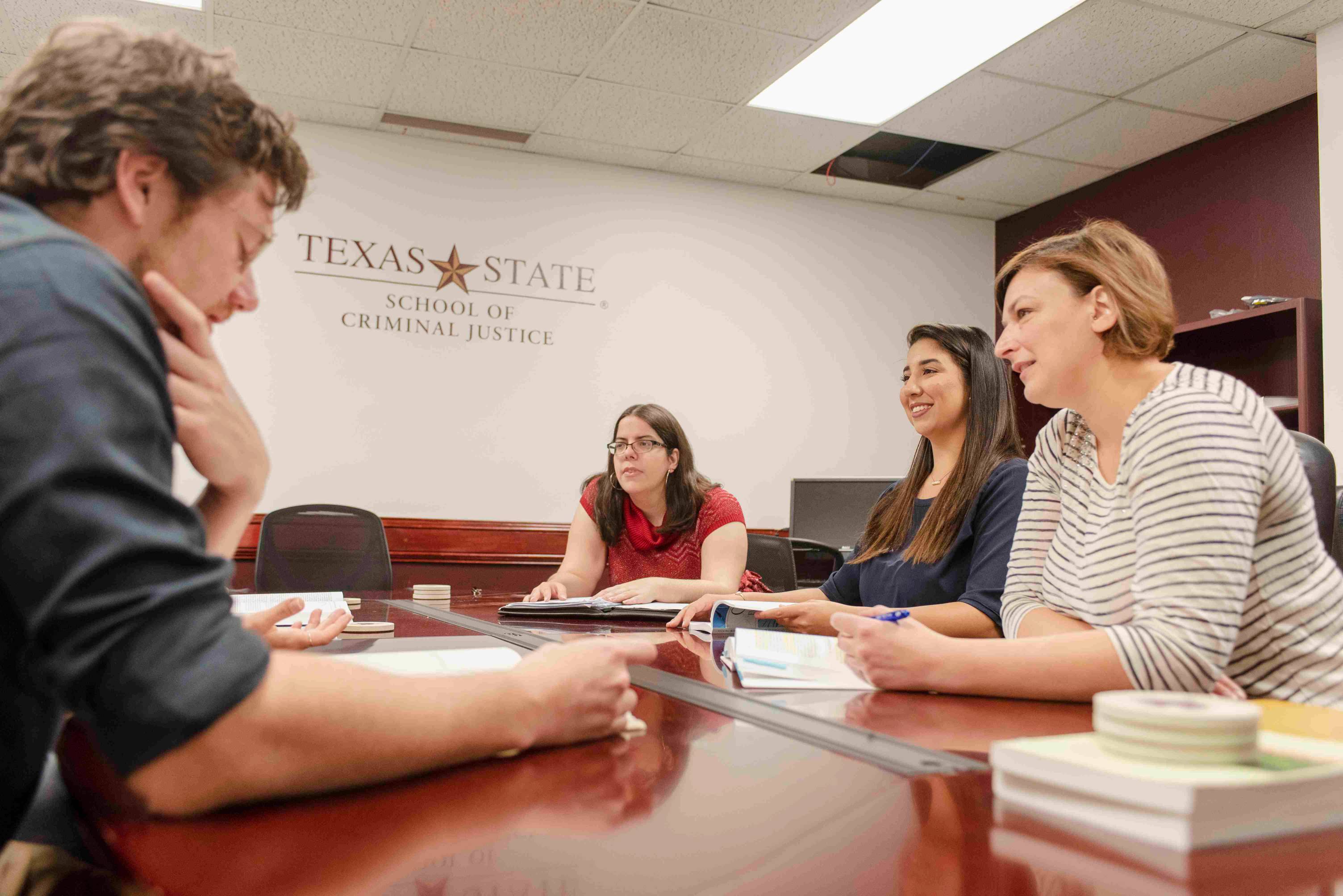 staff and students sit around a table to talk with the texas state university logo on a wall behind them