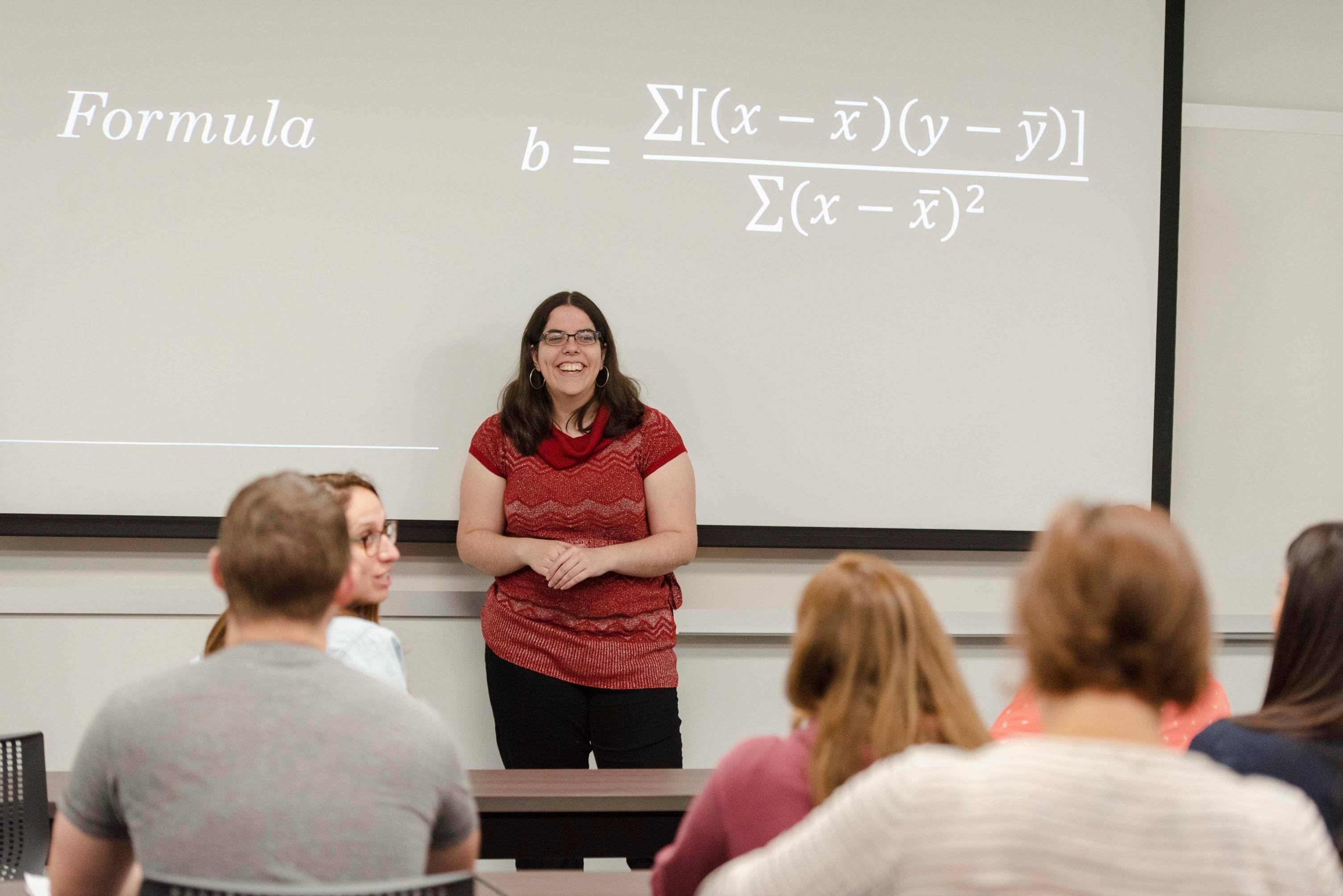 a professor teaches a doctoral class at the front of a classroom with a math formula on the board behind her