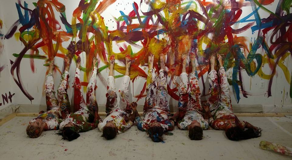 students laying on floor in front of a wall covered in splatters of colorful paint.