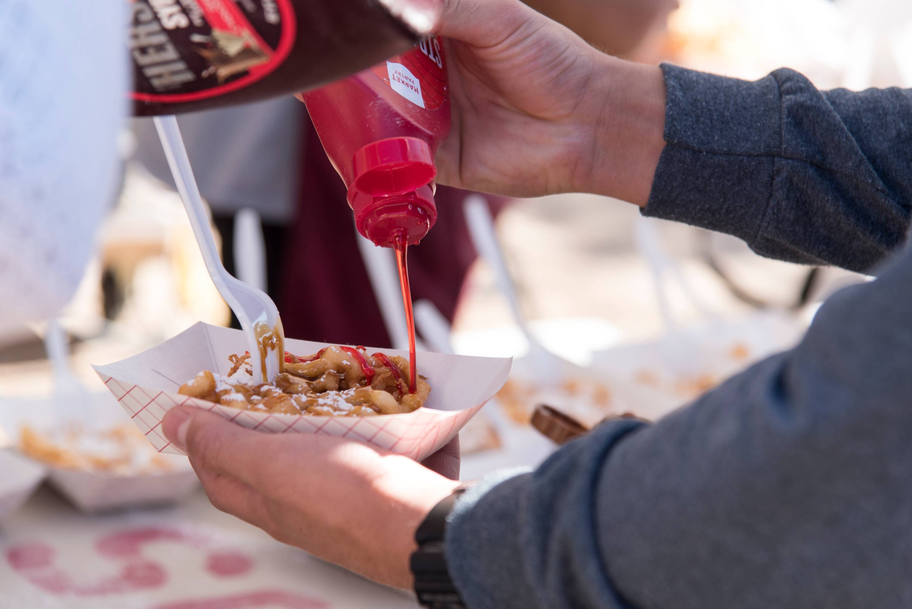 Two hands pictures with one holding a paper plate with a powdered sugar funnel cake while the other hand squirts a red syrup on top of it. 