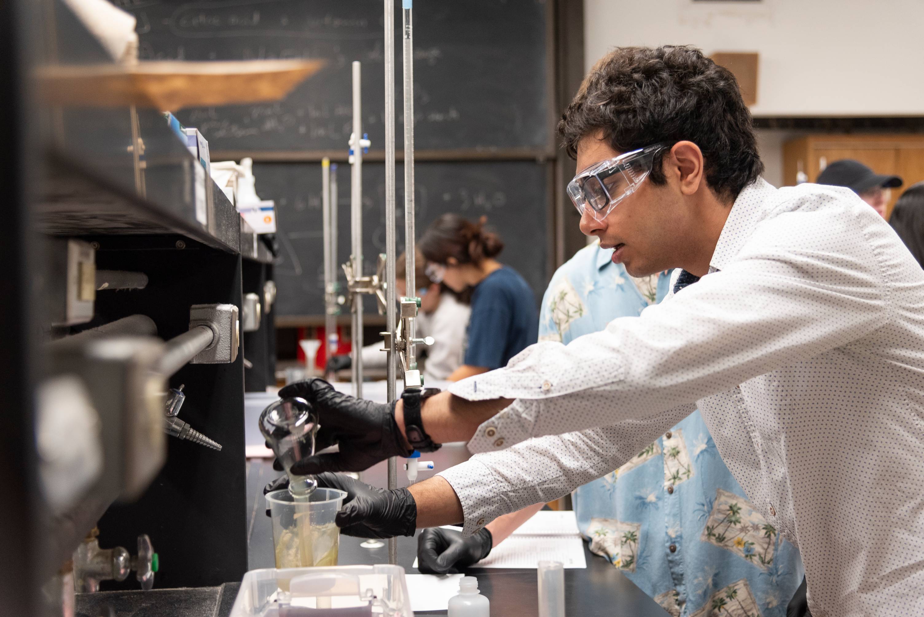 Student uses a flask to transfer liquid to a beaker while other students in the background work in the lab.