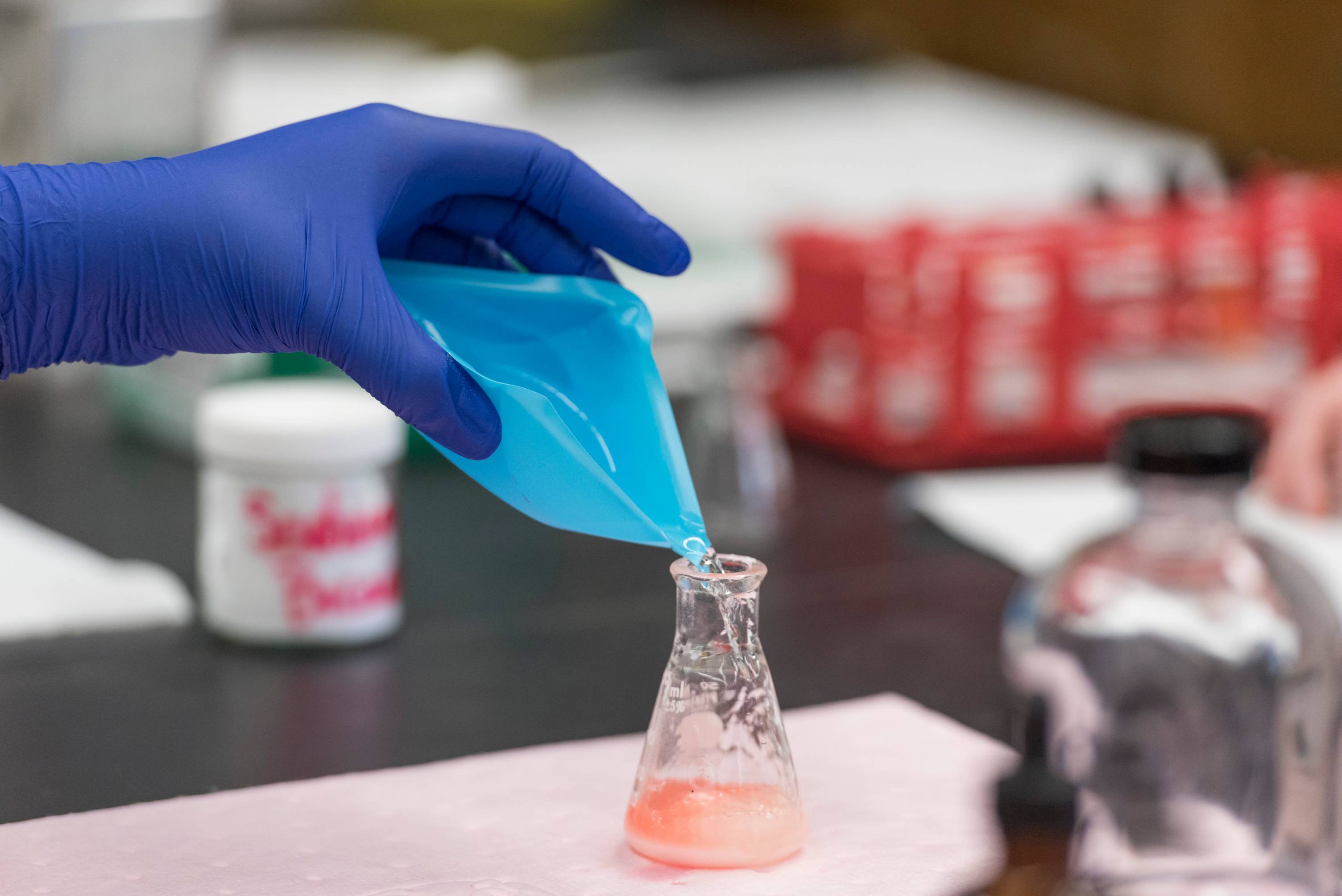 Blue gloved hand pours a clear liquid into a beaker with an orange slurry. 