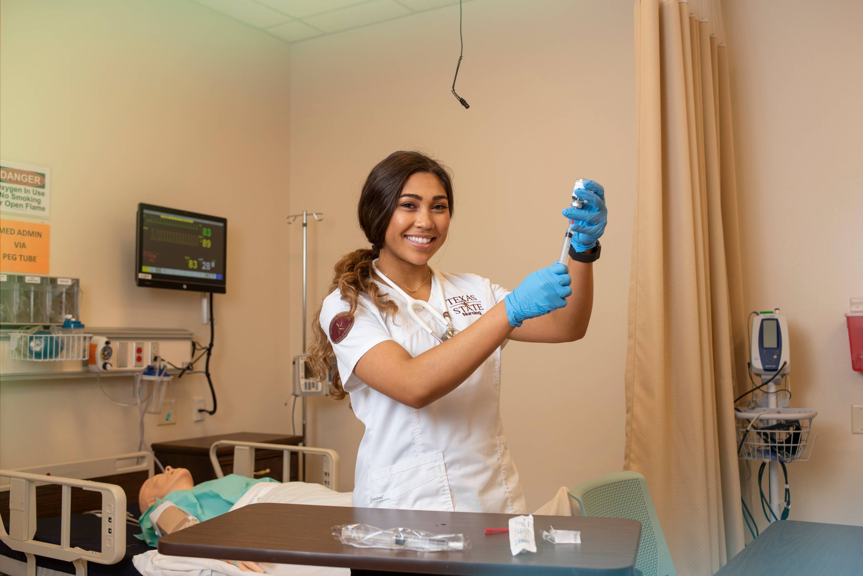 Texas state student in a clinical setting wears scrubs, and blue gloves smiling.