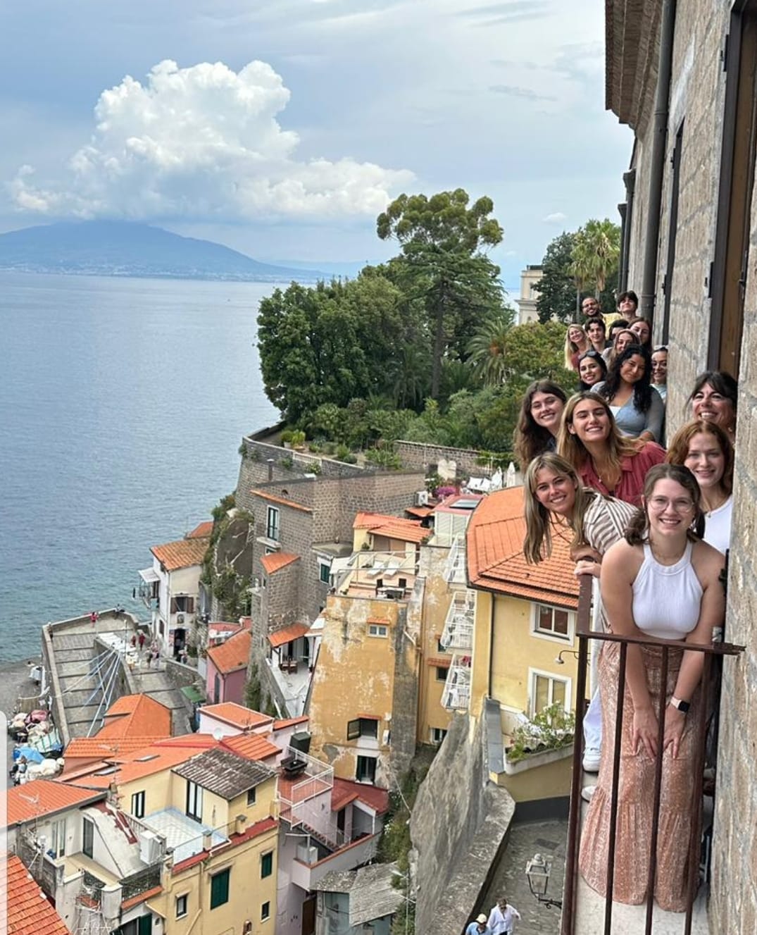group of women and men smiling, hanging out from balconies, Italian city and sea in background