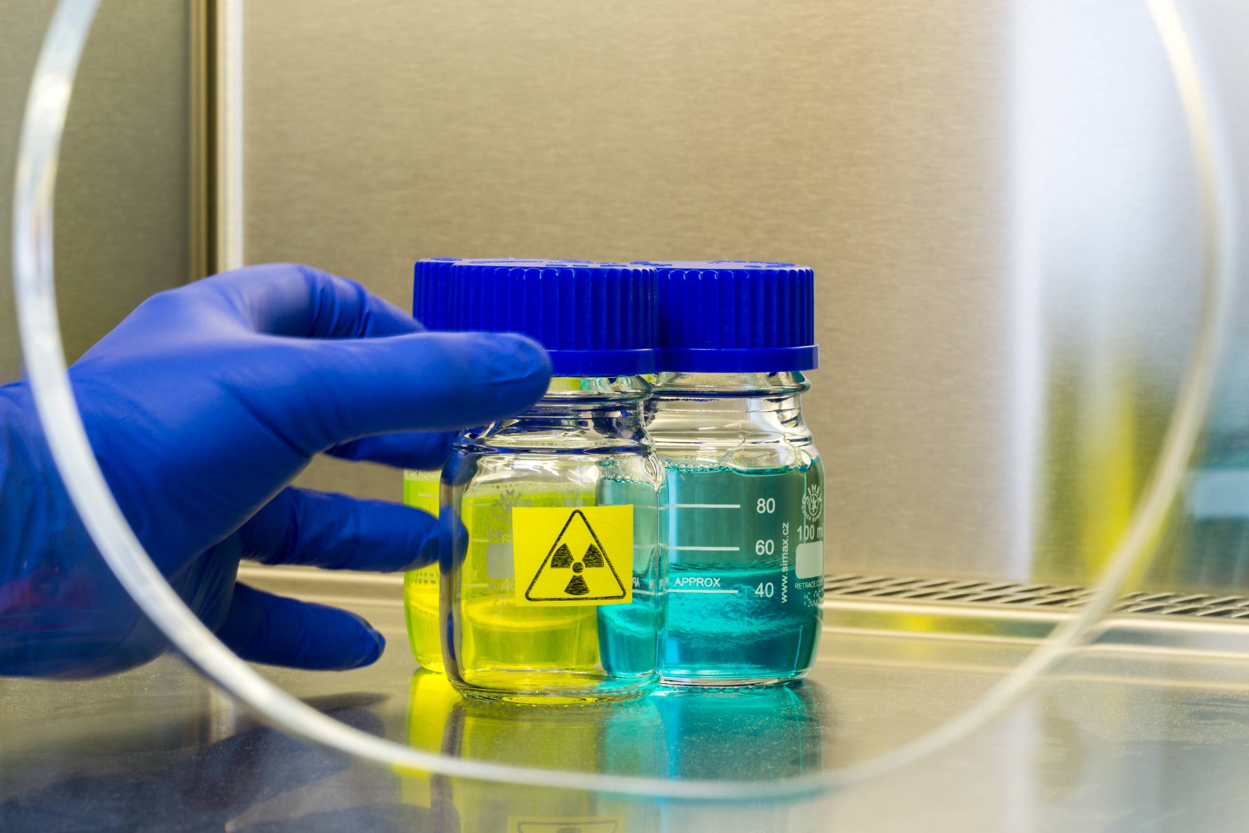 A gloved hand handles 3 vials. The back left vial holds a yellow liquid, the back right a blue liquid, and the front center a clear liquid with a radioactive symbol. 