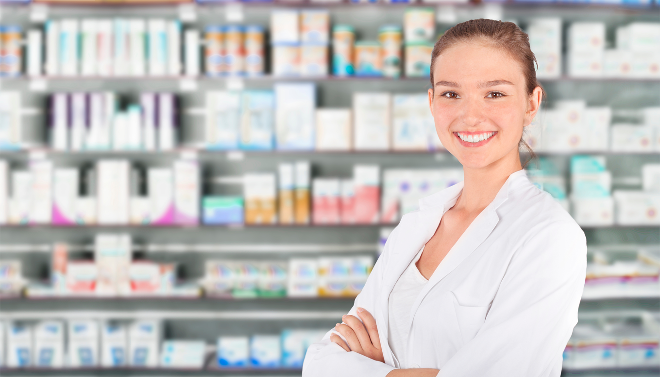Female pharmacy technician standing in front of a shelf with medicine on it.  