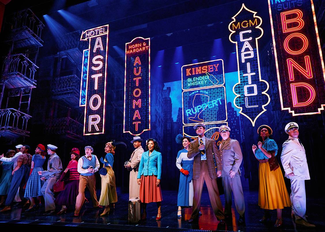 broadway play cast member on stage with multiple lighted signs in background