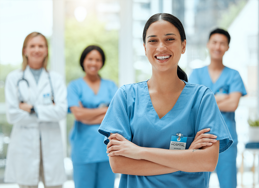 Female medical assistant in slacks standing in front of other medical workers.