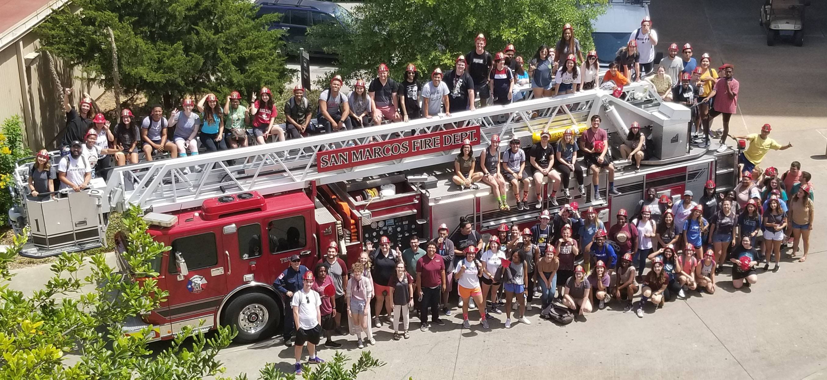 A large groups of students pose in front of a fire trucks with firefighters. 