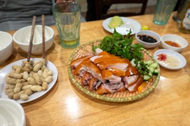Picture of slices of roasted pig on a plate in the center of the picture, with a plate of peanuts in the shell on the left, three small bowls of dipping sauces on the right, and a green glass with one inch of beer in it at the top. .