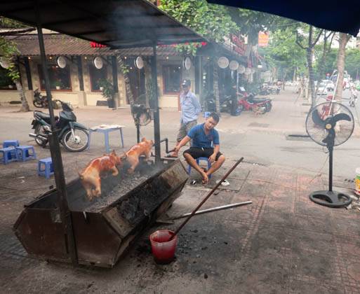 Picture of two full pigs on a spit being turned over a charcoal brazier and a seated man turning the spit. There are two fans blowing on the man and smoke coming from the brazier.