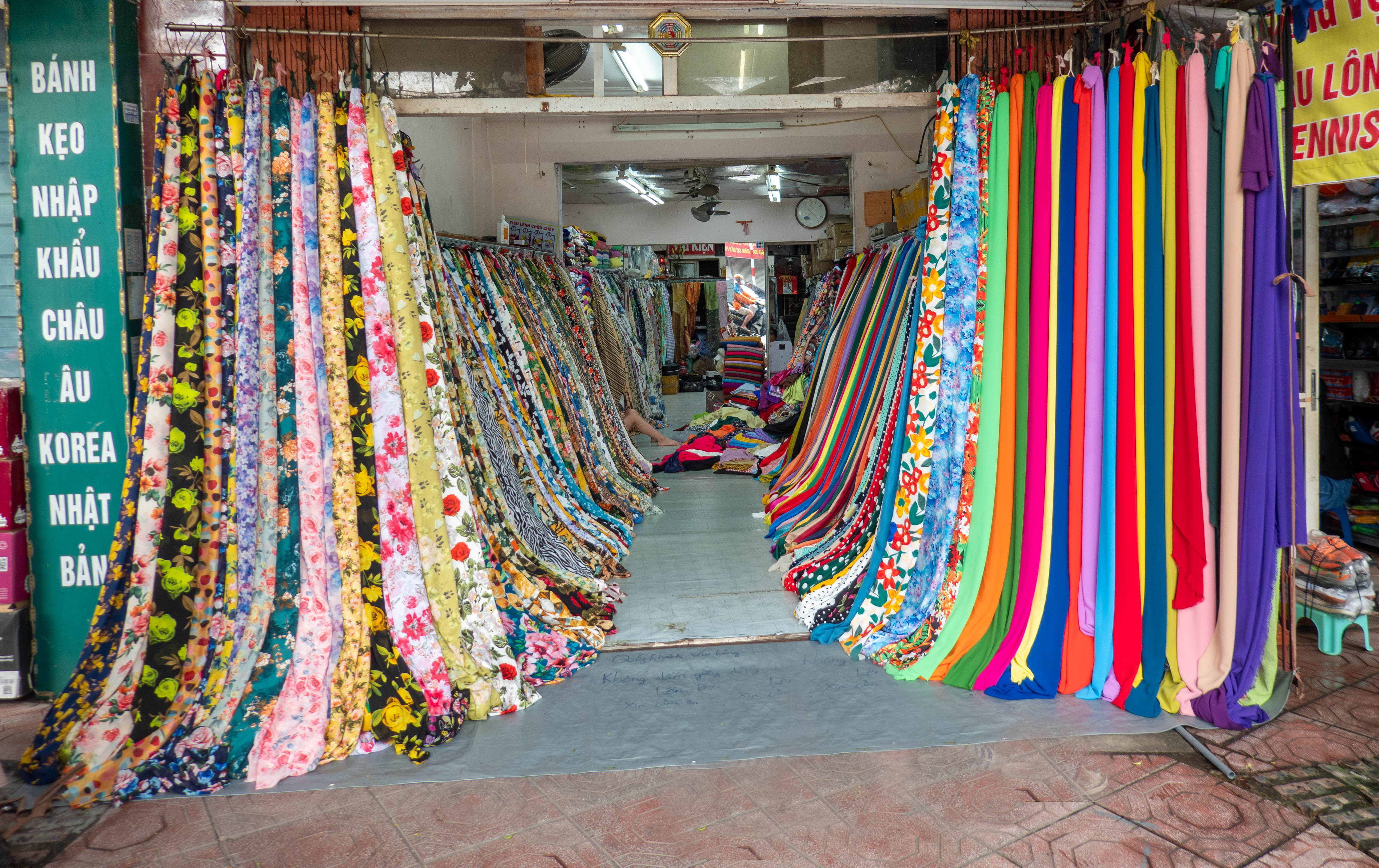 Picture of looking into a fabric shop with vertically hanging multi-patterned, colored fabric samples on left side of entrance hanging from the entrance back into the back of the store and solid colored samples on right side doing the same. Three quarters of the way back into the shop on the left side there is a person sitting among the samples but all you can see is the leg sticking out.