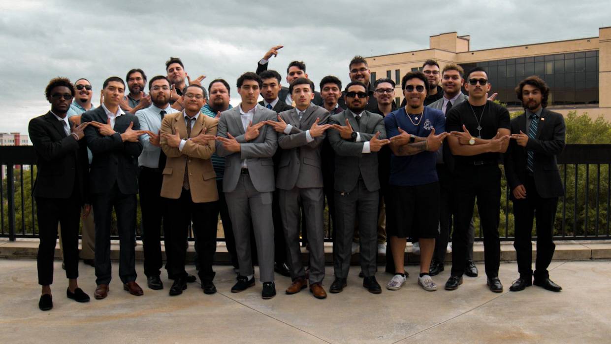Large group of Sigma Lambda Beta men holding up their hand signs outside on the LBJSC balcony