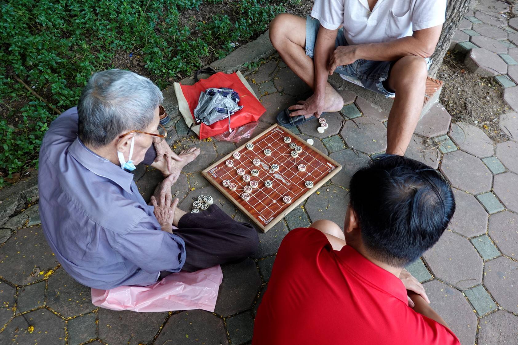 Three men sitting on paving stone walk. Two are playing a board game and the third man is watching them play.en watching board game in park
