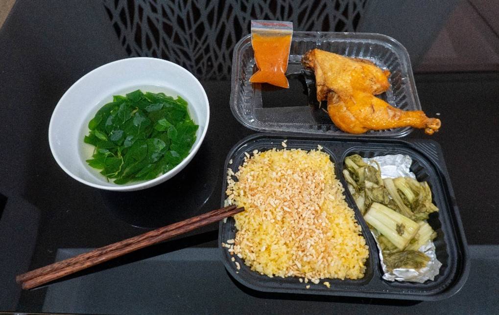 Picture of table top with a bowl of green leaves in broth, chopsticks andto the right a plastic container of rice and pickled green lettuce-like vegetable with the lid directly above it on the table holding a chicken thigh and drumstic.
