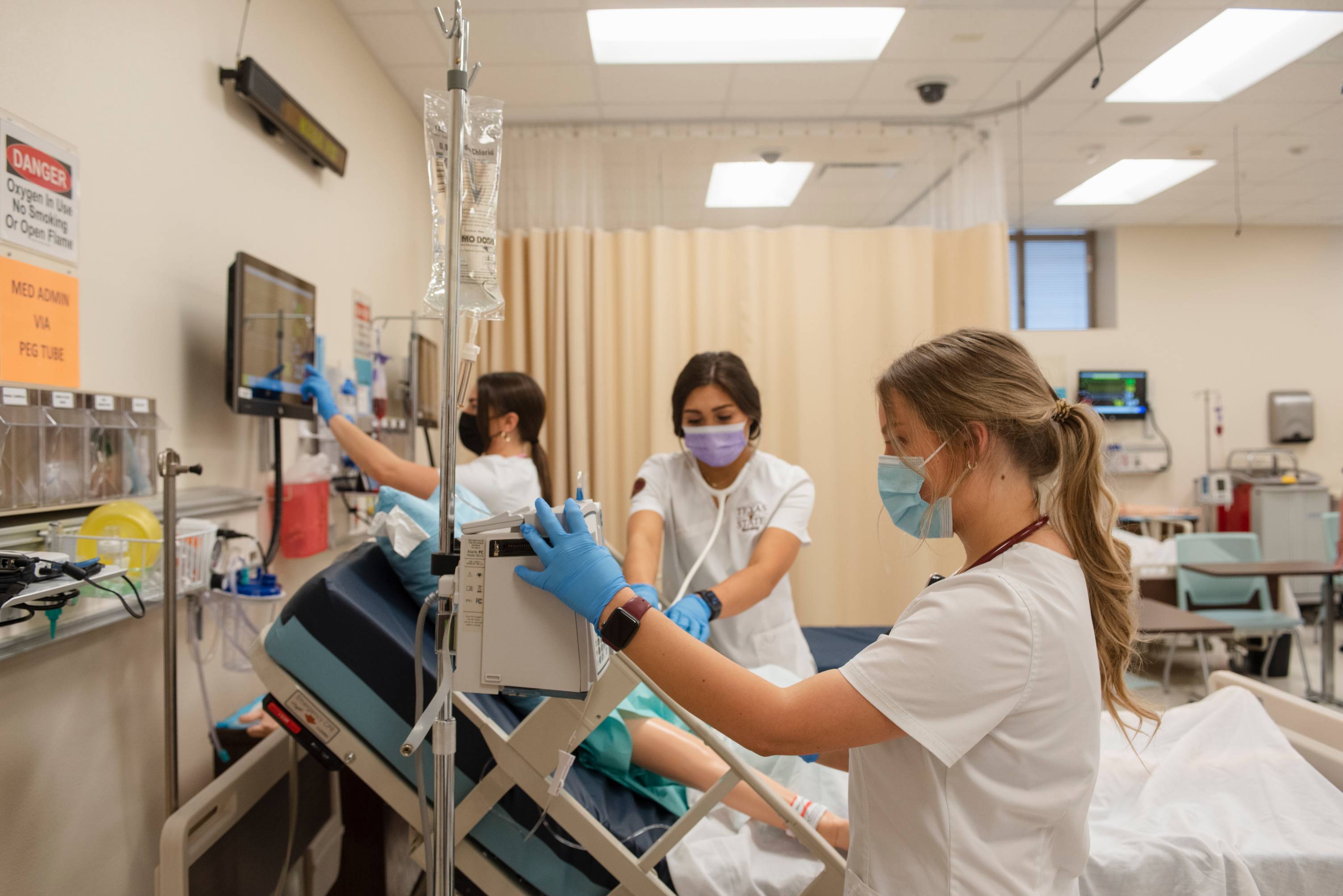 Three students working around one hospital bed occupied by a manikin in the Nursing Simulation Lab