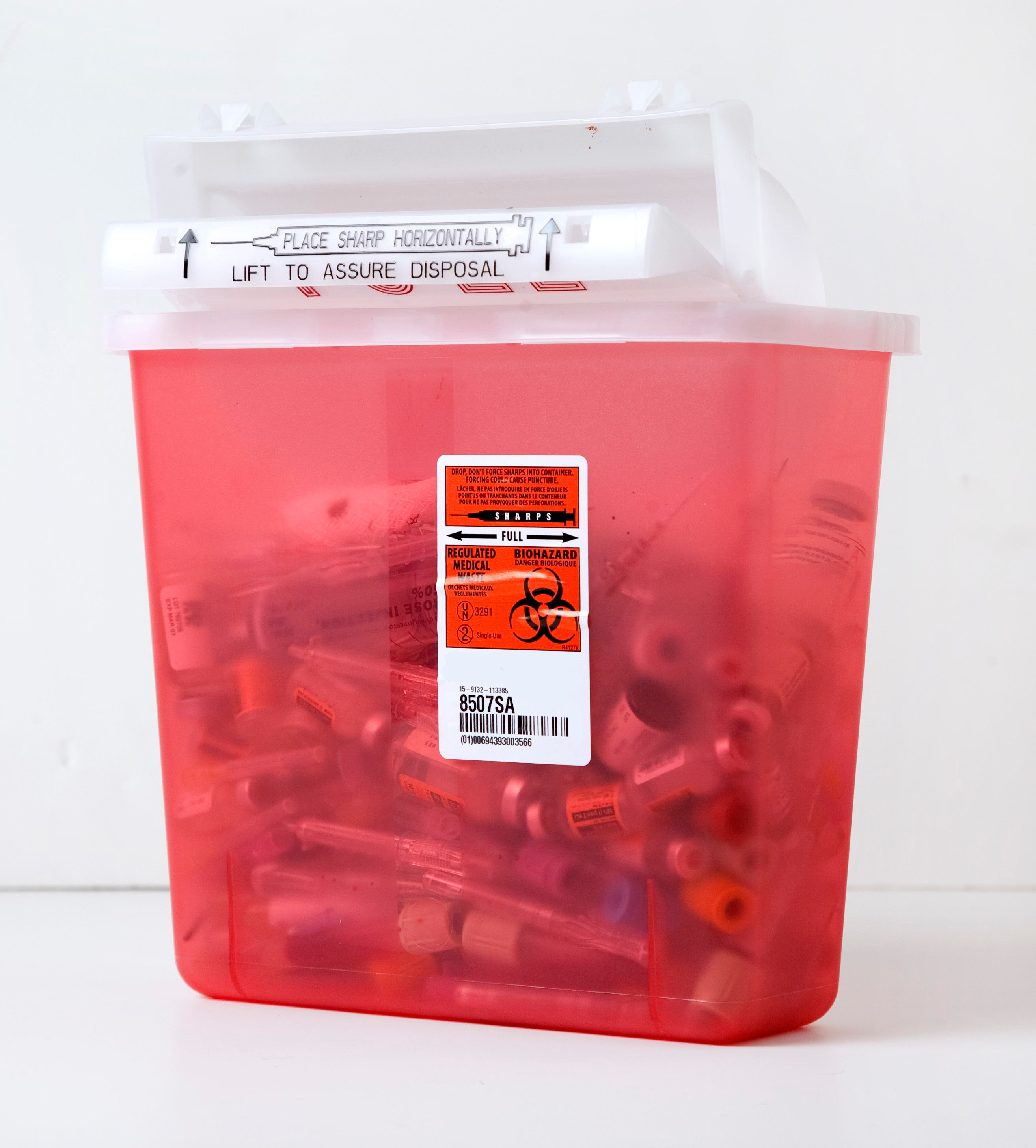 A red translucent box with a clear lid that is halfway full with needles and other types of sharps. 