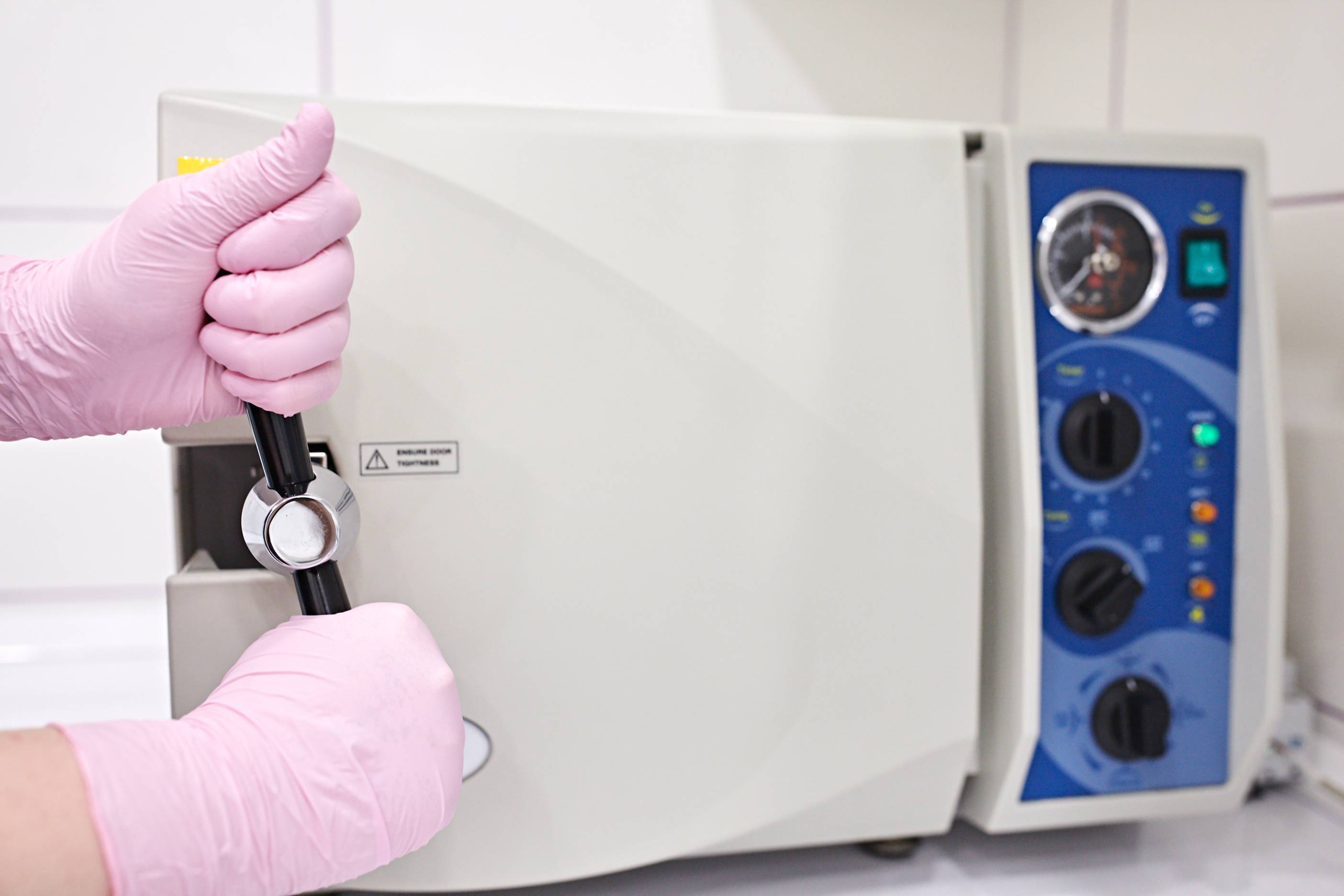 Two pink gloved hands twist a handle on an autoclave machine to open the door. The machine is rectangular with a white door and blue knobs on the right. 