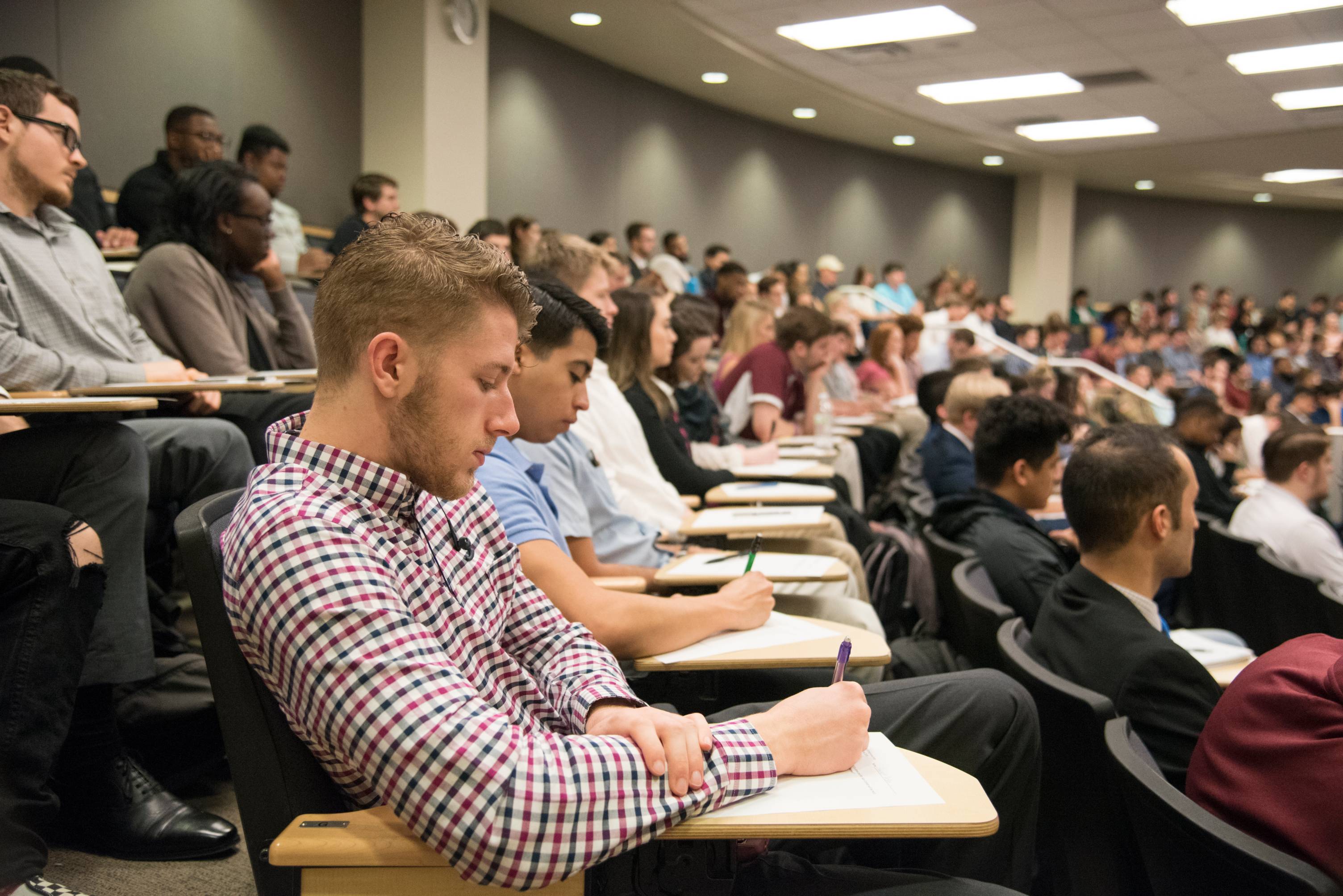 A fully-occupied lecture hall with stadium seating, students look concentrated on listening and taking notes