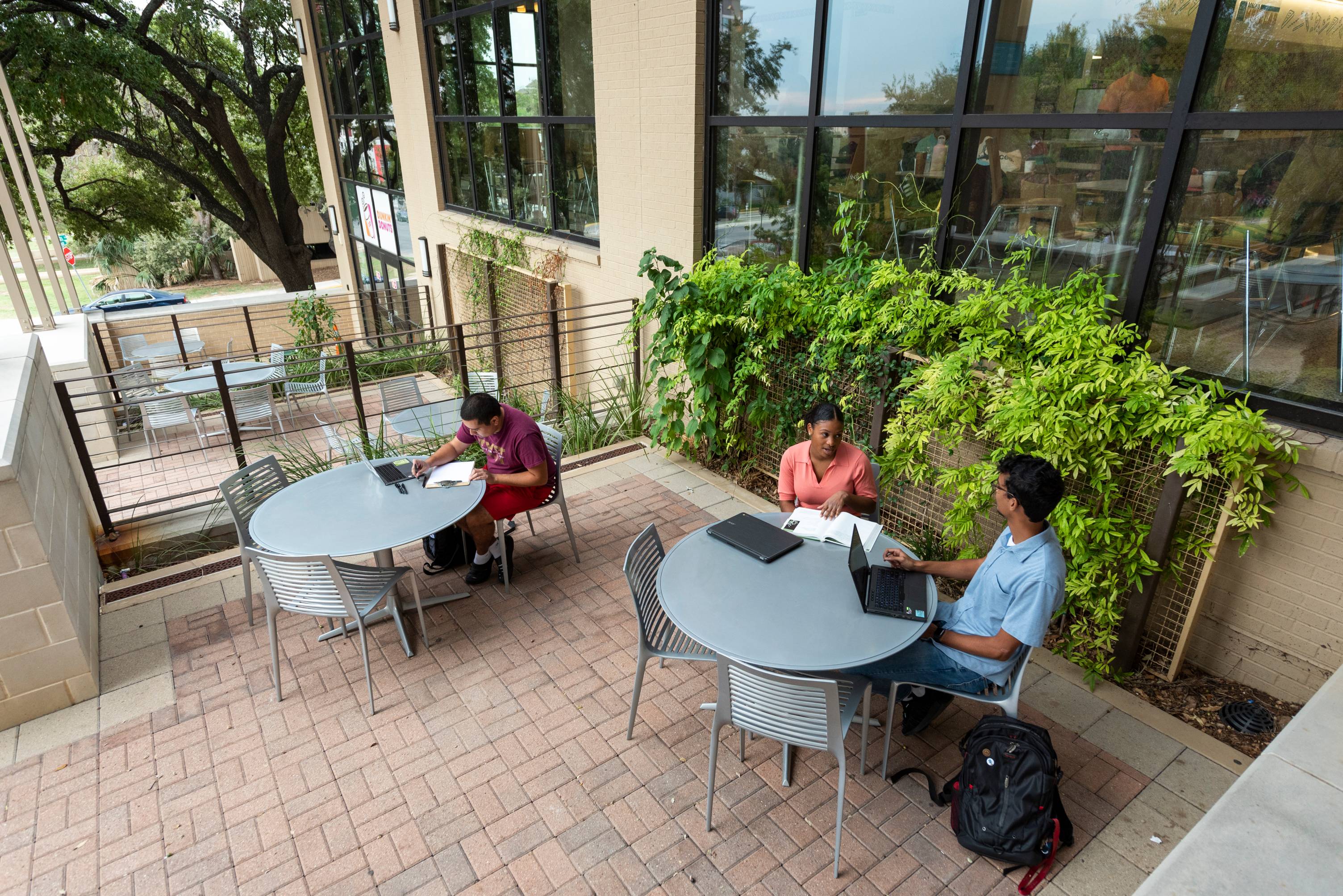 Three students sitting at tables outside of Jones Dining Center working on schoolwork with their laptops and textbooks out