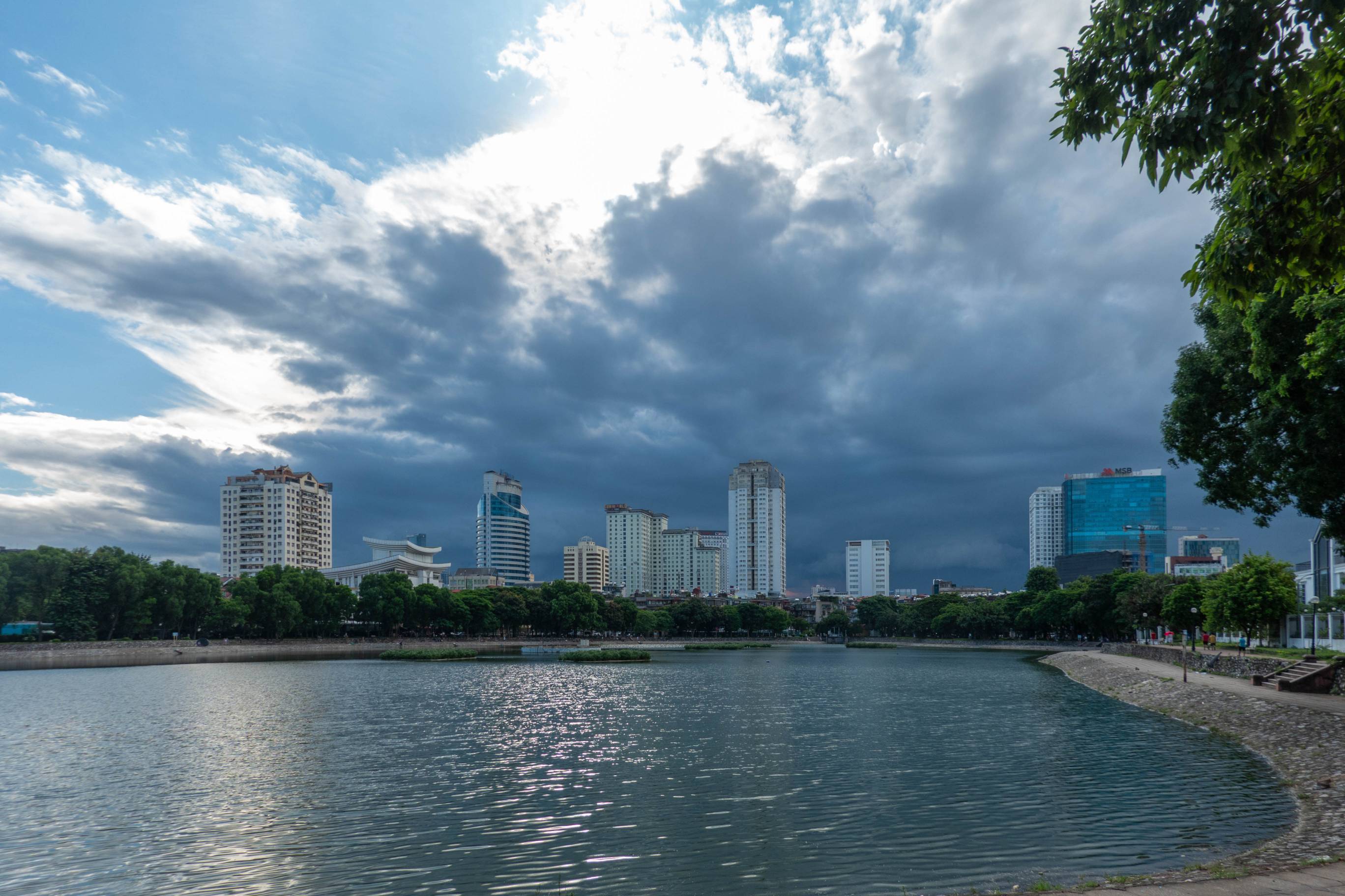 picture of lake with tall buildings on far side of lake and dense rainclouds moving in over buildings toward lake.