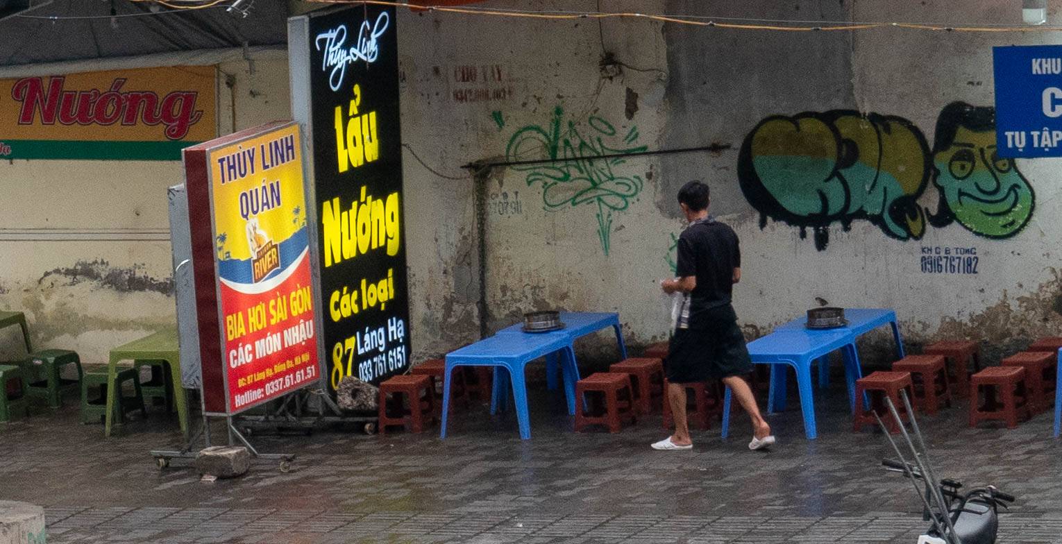 Picture of many walking in front of two blue tables iwth brown stools that have a hot pot on them. There is spray painted graffiti on on the wall to the right of the man that has letters and a face.