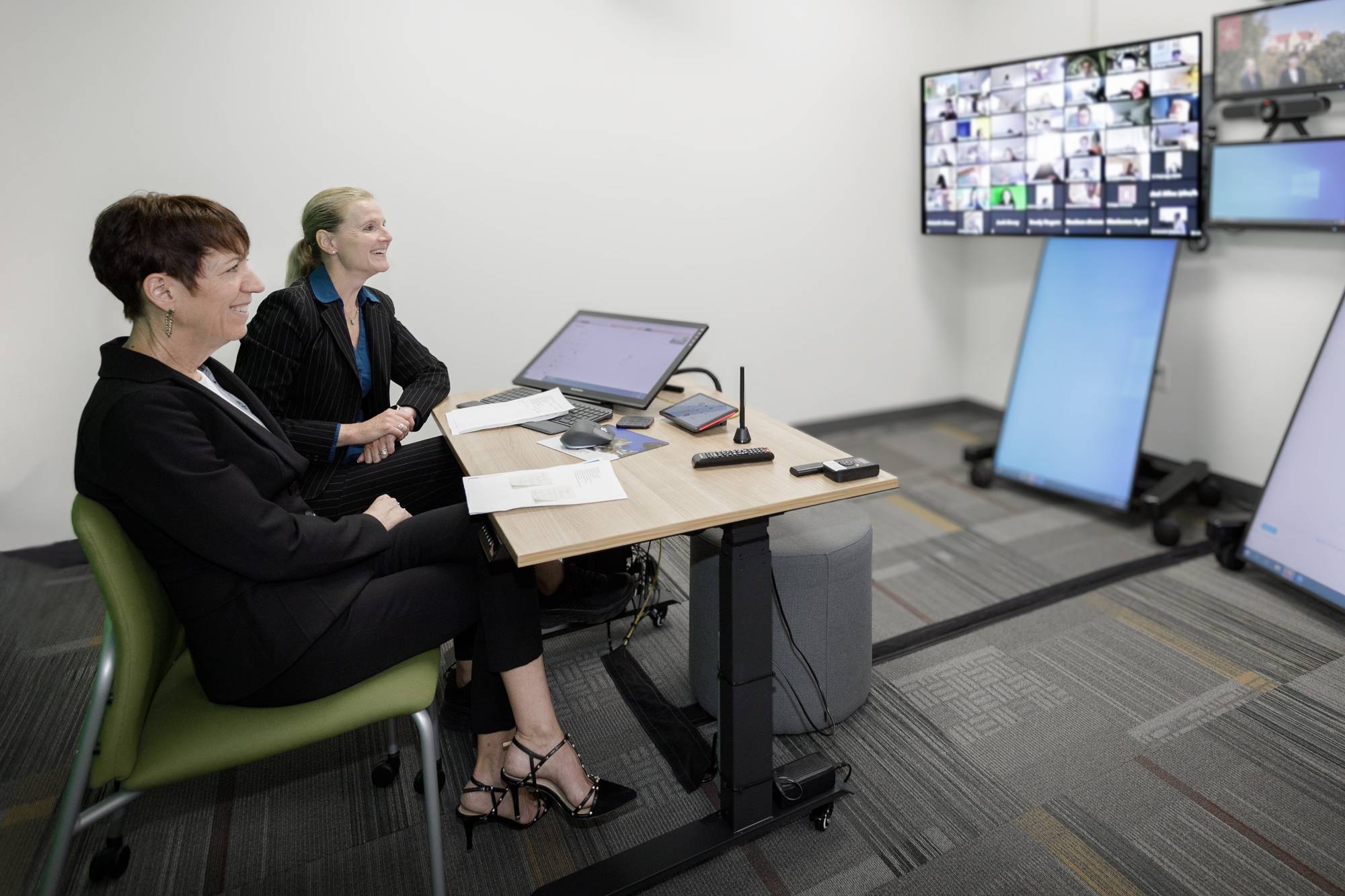 two women sitting in room looking at large group of people in virtual meeting on computer screen