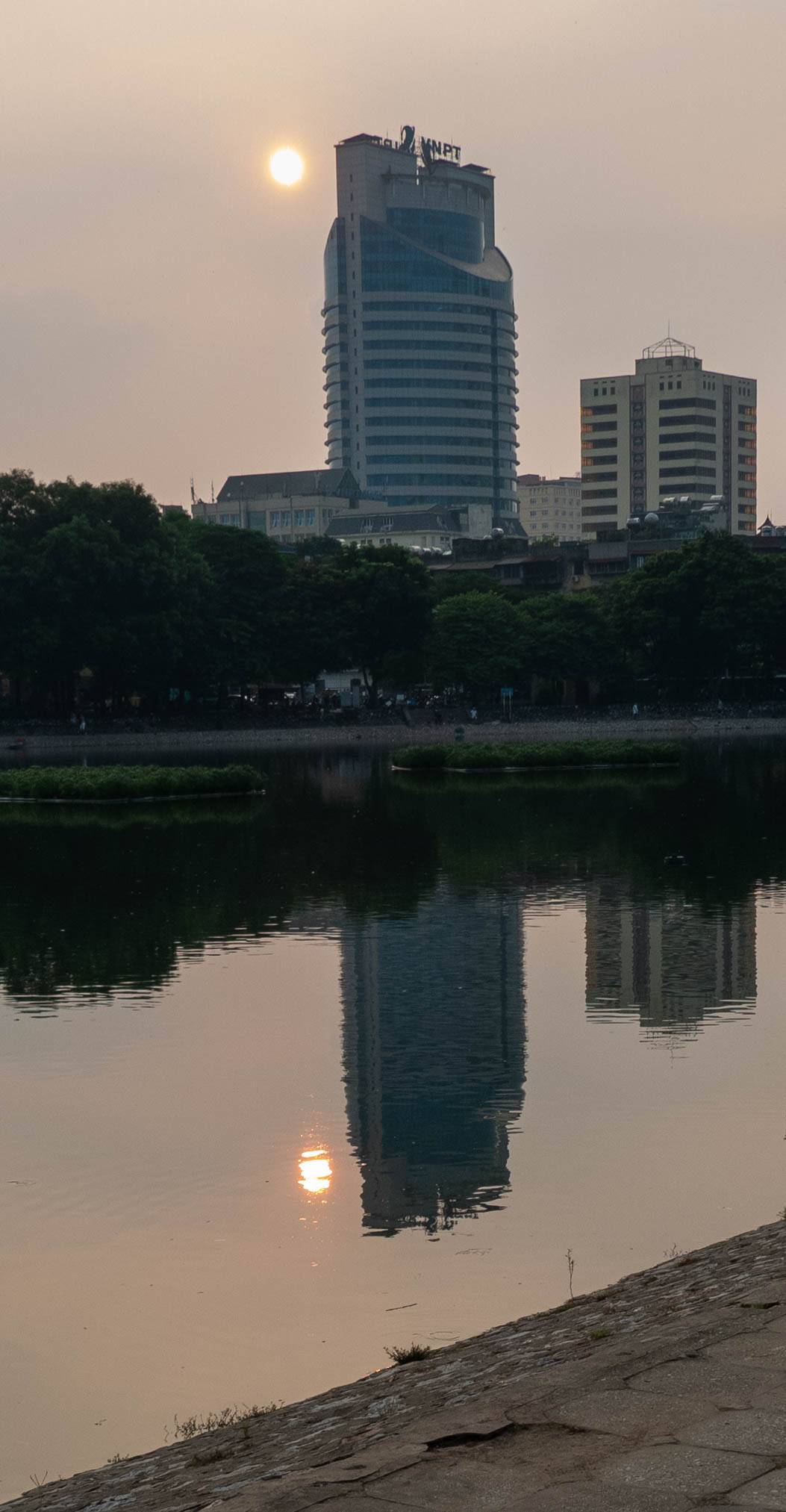 Building and sun reflecting in calm lake. Sun is to the left of the tall building.