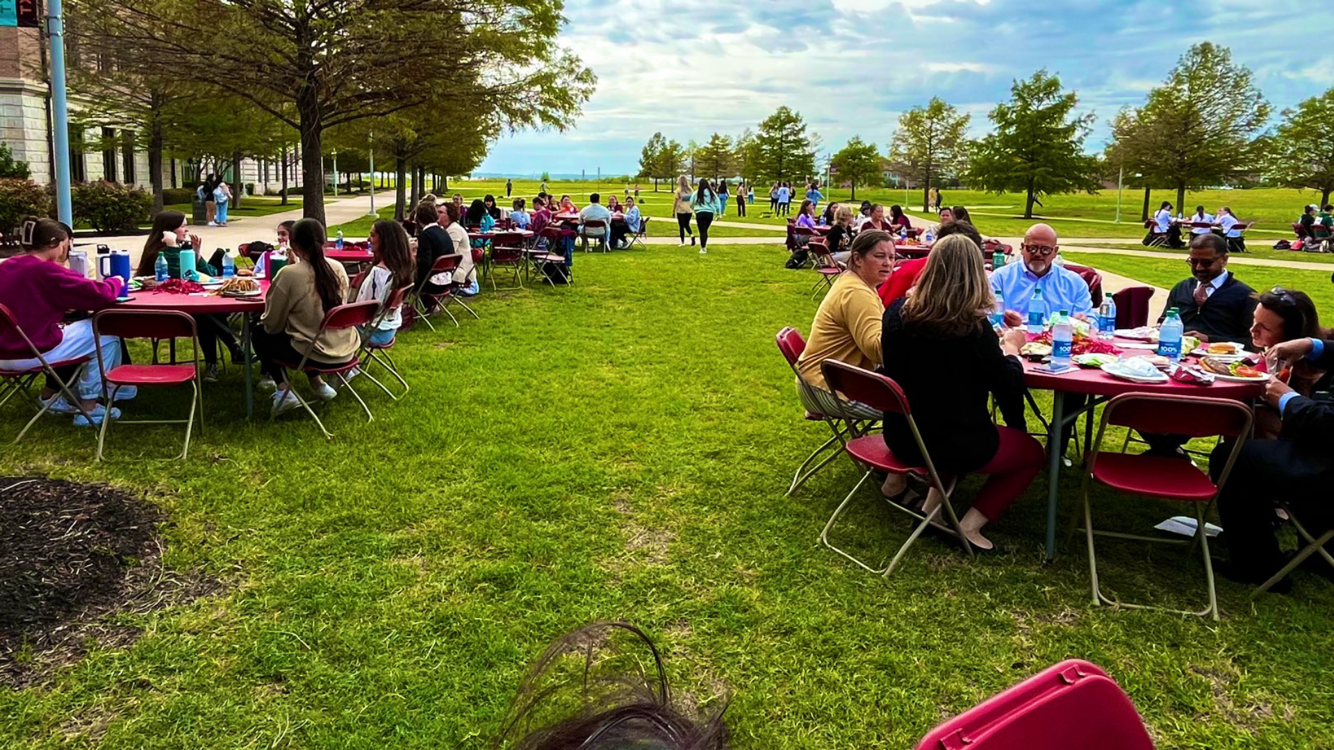 Groups of students, staff, and faculty sit at tables spread out across a grassy area.