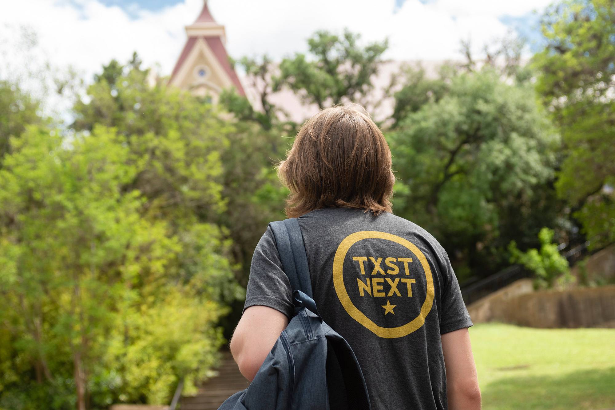 Student with TXST Next shirt walking towards Old Main holding a backpack