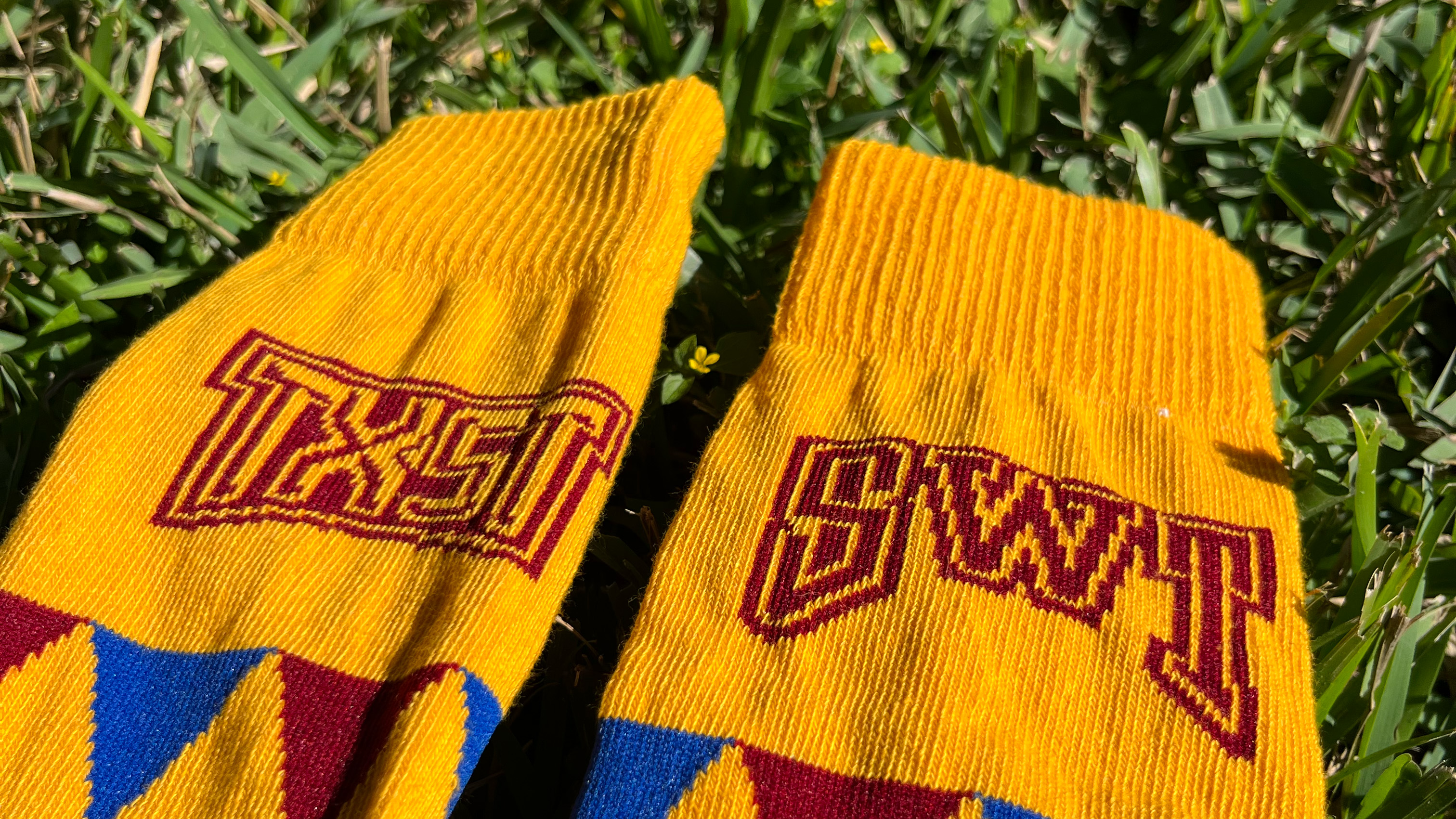 Yellow gold socks with maroon and blue argyle print and TXST logo on one sock and SWT logo on the other sock laying in the grass