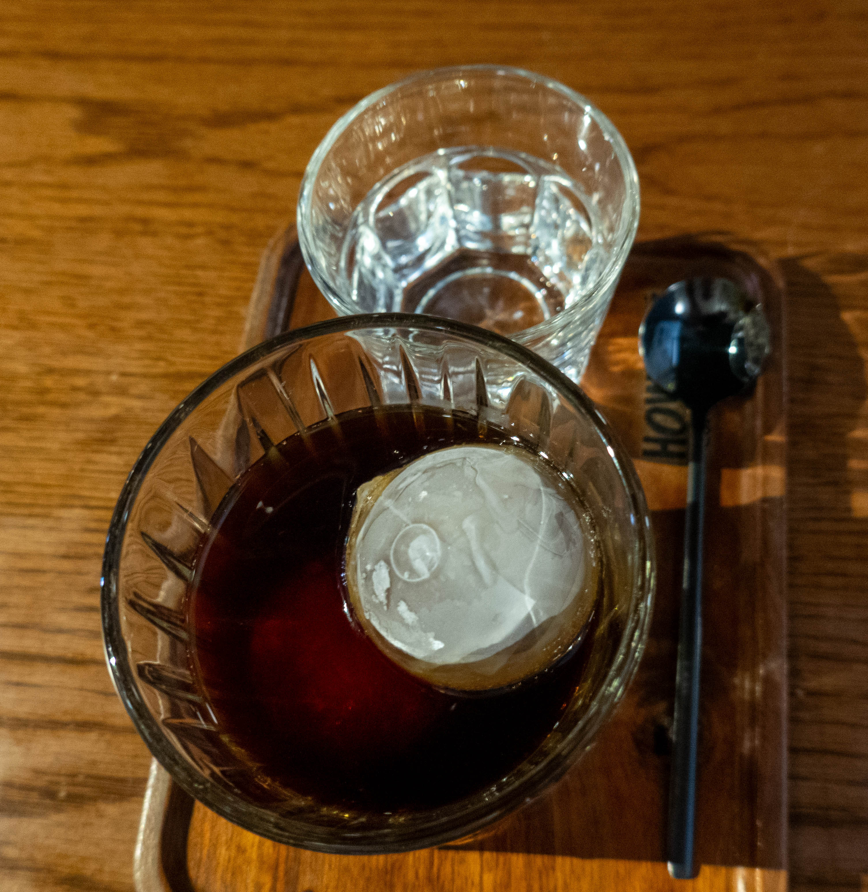 Picture of glass of black coffee with a round icecube that has a round dot at nine o'clock. Above galss is shot glass of water and spoon to right. Items sit on a wood serving tray on a wood table.