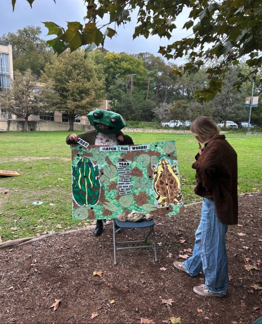 A TXST student reads a poster with a person dressed as a frog behind it.