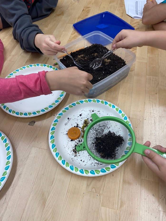 Students use sifters and spoons to uncover artifacts in a dig box.