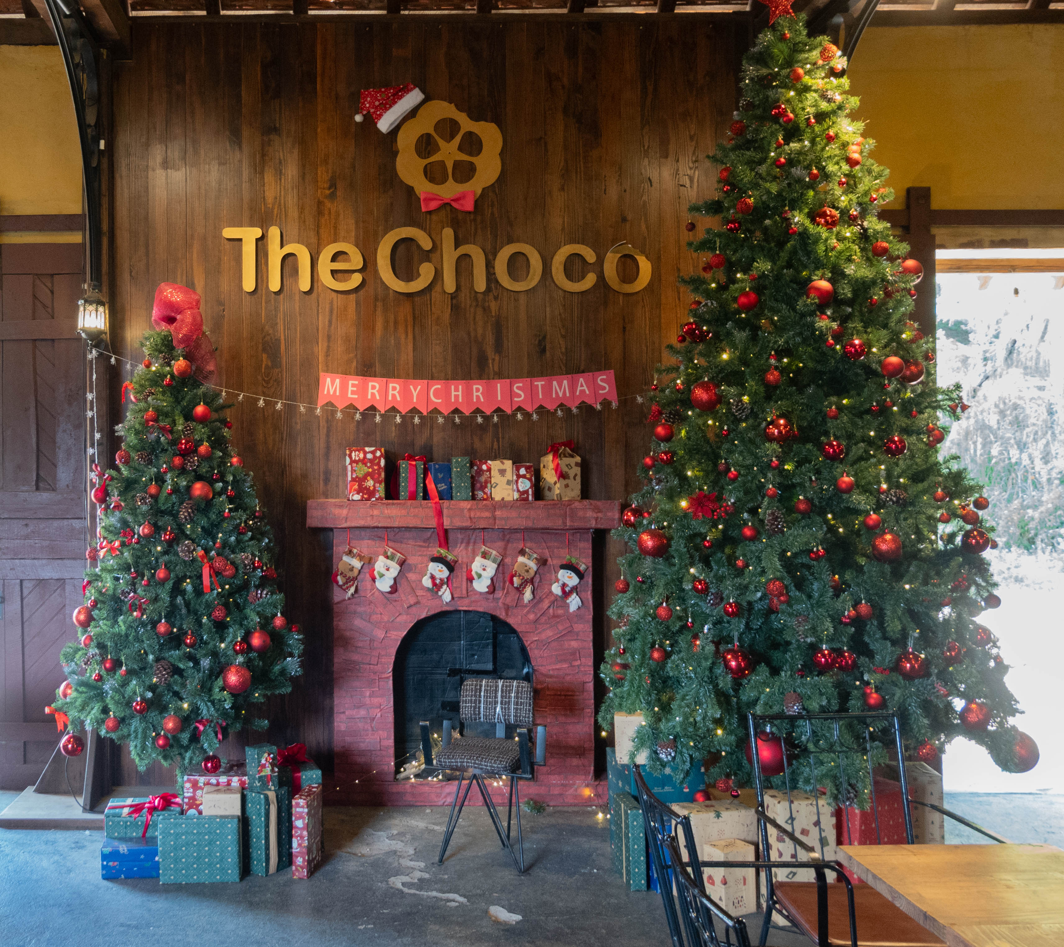 In the center fo the picture is the name "The Choco" under a symbol wearing a Santa Hat pointing to the left. Underneath the name The Chaco is a banner saying Merry Christmas. Under that is a fireplace with stockings hung on the mantle and to the left of the fireplace is a medium sized decorated fake Chritmas tree and a larger one is to the right of the fireplace. There is a chair in front of the fireplace.