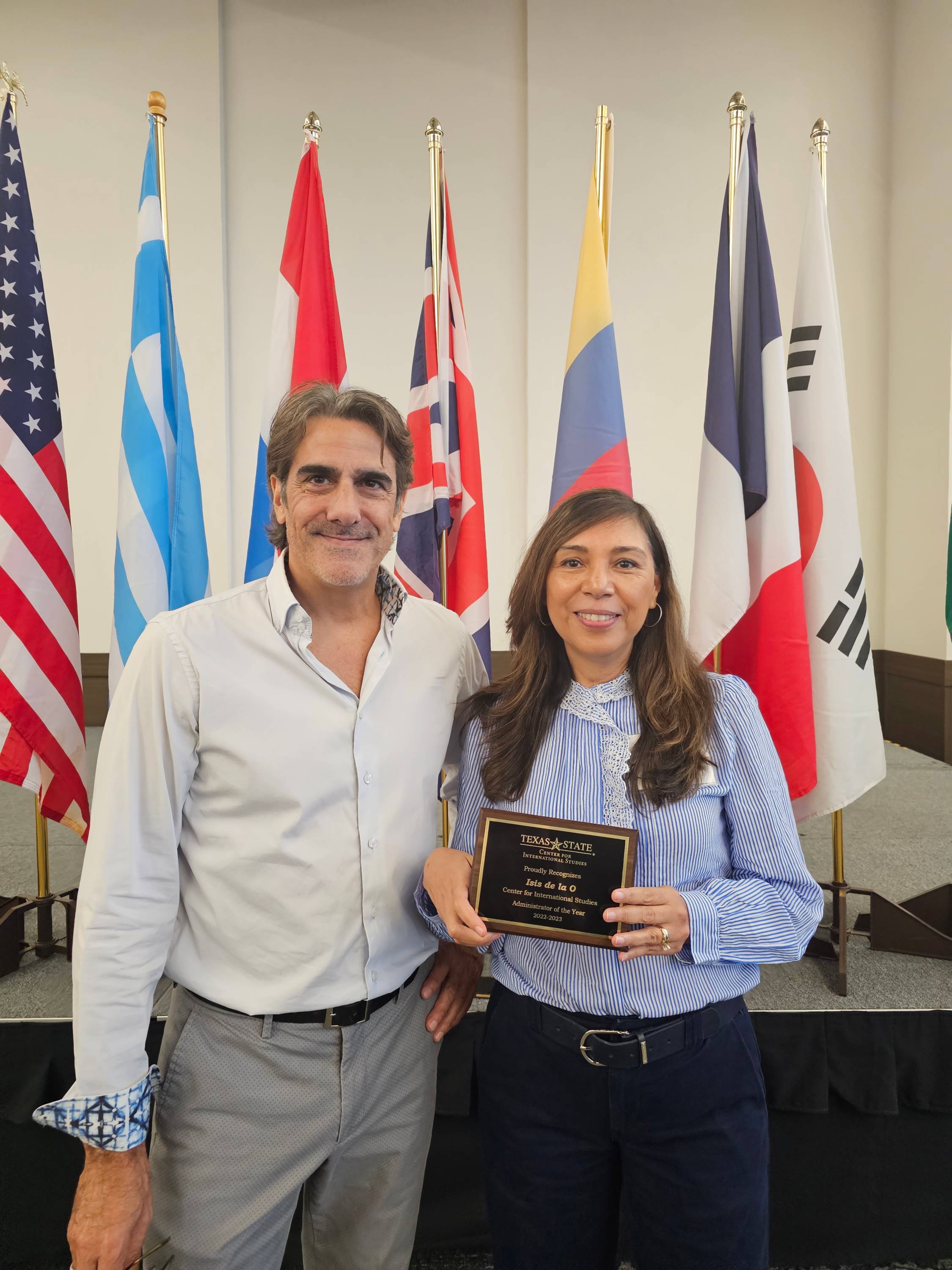 Dr. Paul Hart and Isis De La O stand in front of national flags with an award plaque