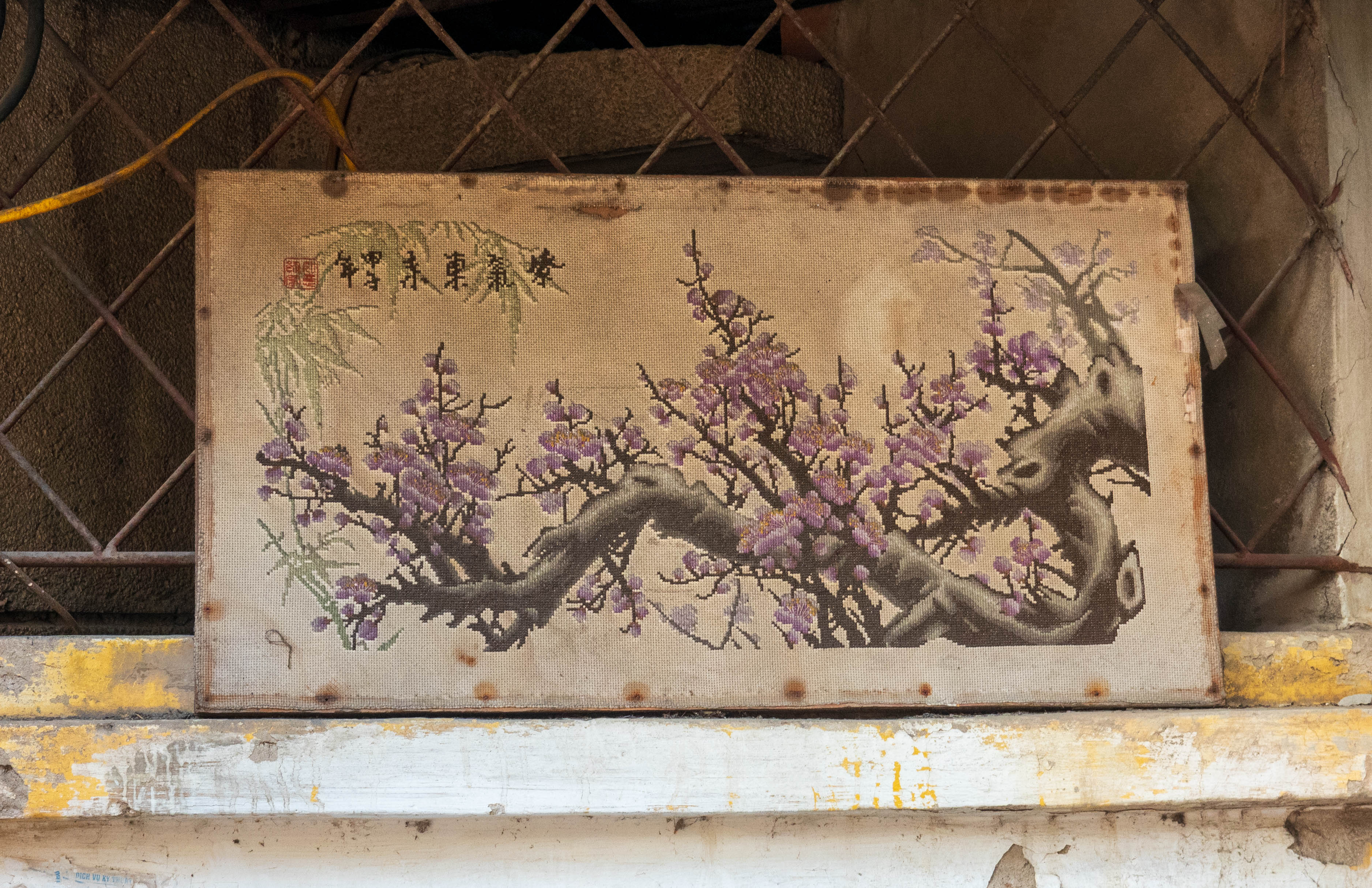 Close-up picture of purple flowers on a tree branch is doen in needlepoint. There is what appears to be Chinese characters and a read seal in upper left hand corner.