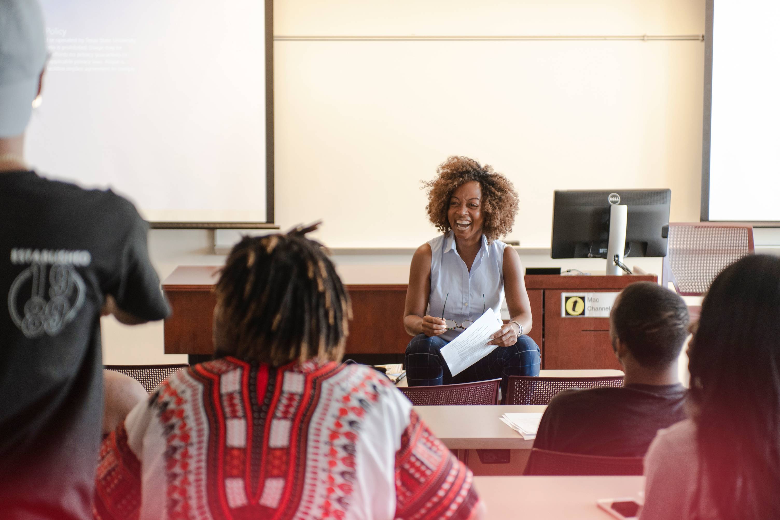 An instructor sits in the front of the classroom with a big smile on her face as she lectures students.