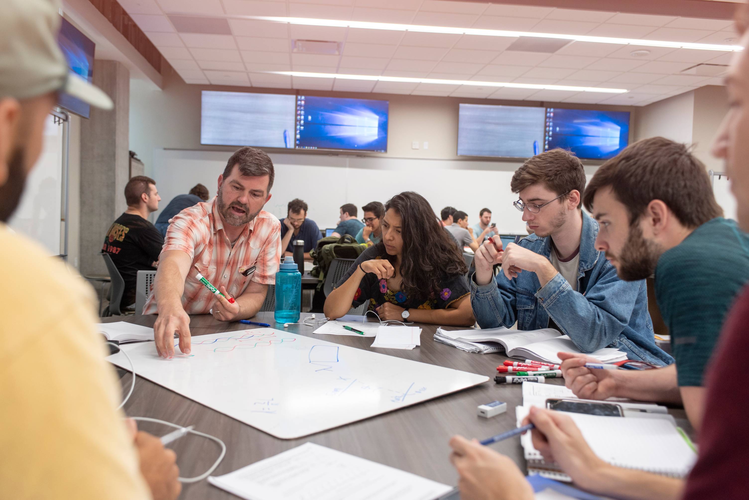 An instructor and three students are gathered around a table with books, notes, and markers. The instructor is explaining a concept and the students are listening.