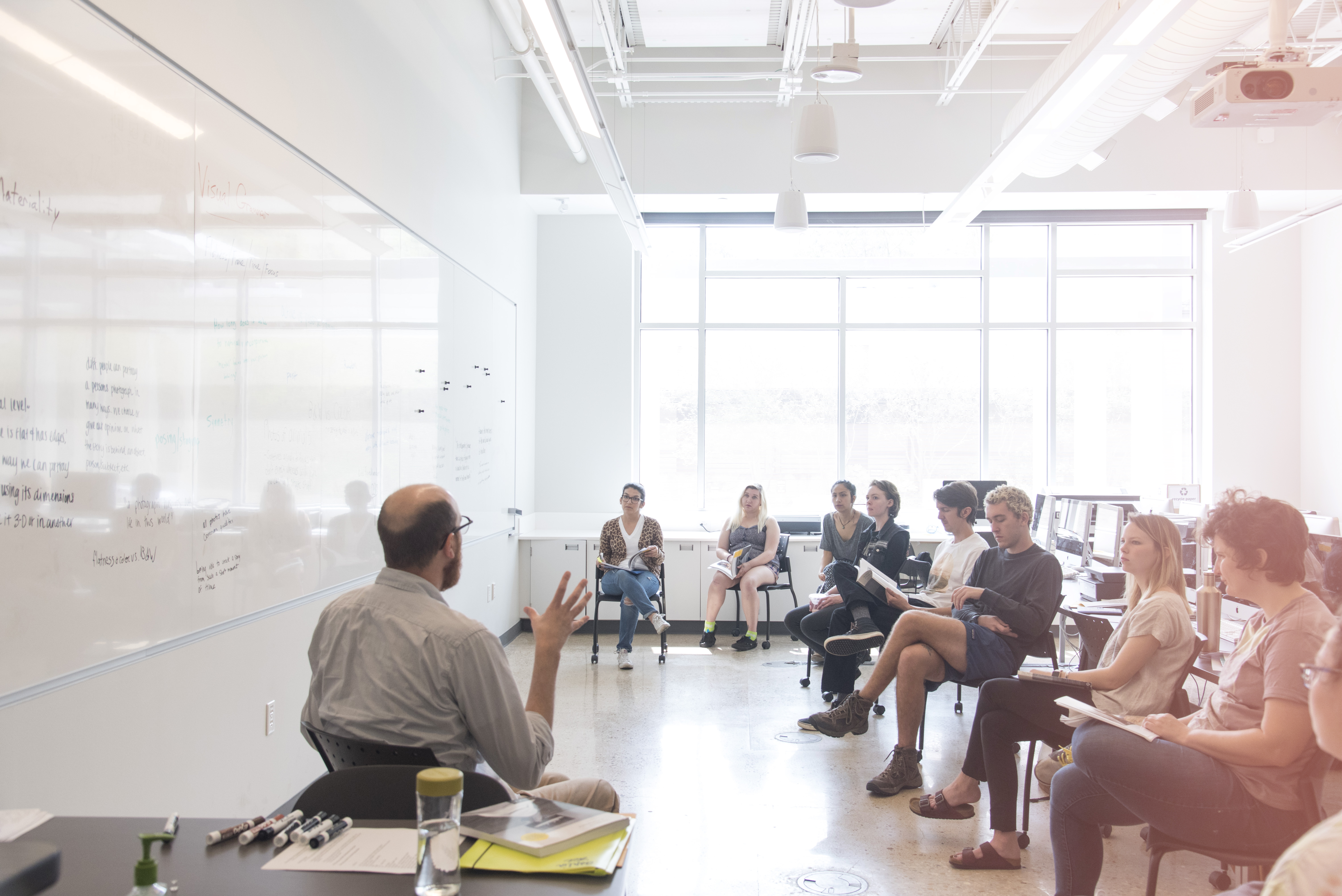 An instructor and group of students sit in a classroom. The instructor is discussing a concept.