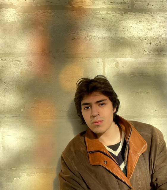 man wearing jacket, open to a v-neck sweater, against light brick wall, shadows cast across entire image