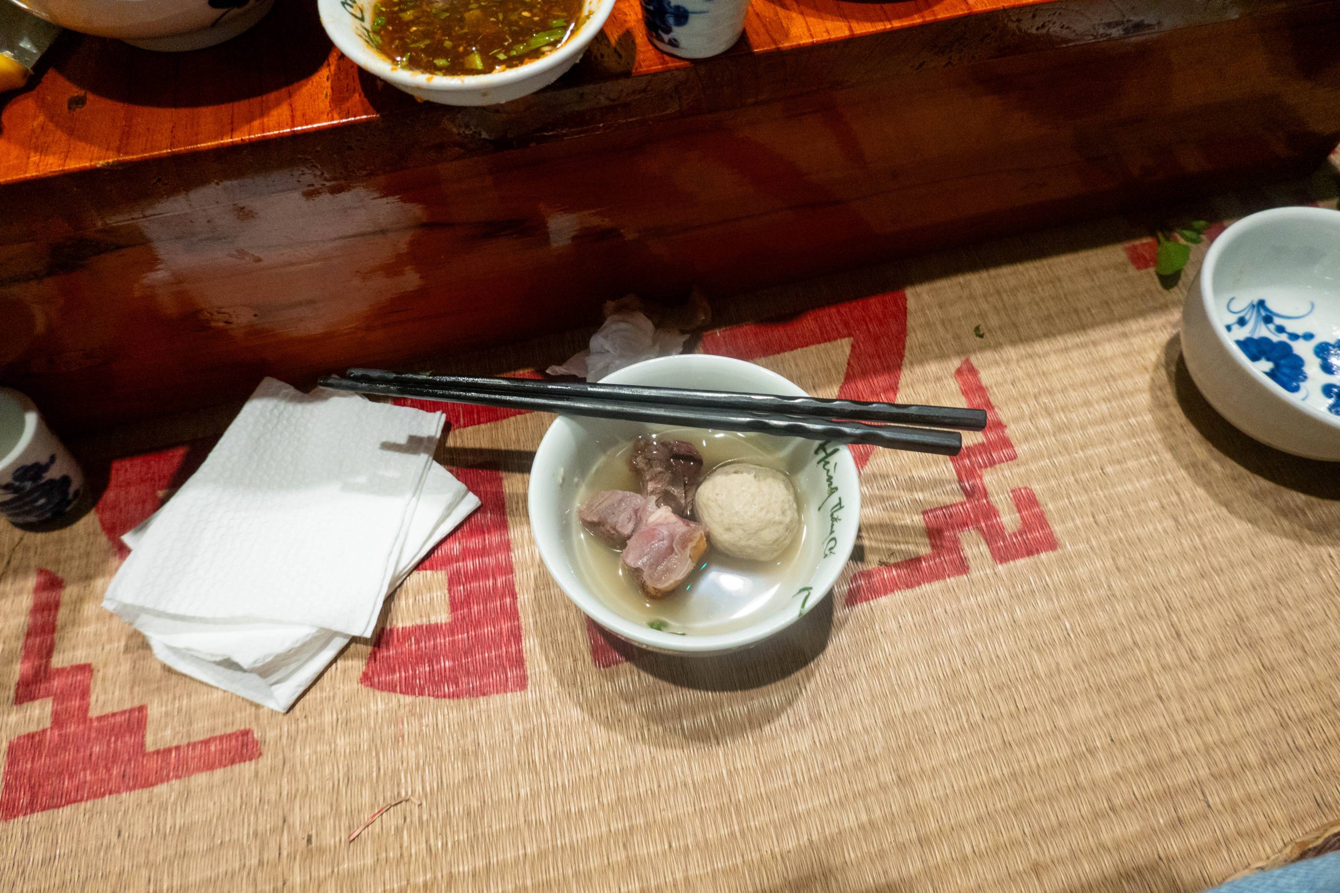 Picture of a bowl with meat and dumping with chopstick laying across bowl at top of bowl. White napkins to the left and a blue and white bowl to ther right are also visible.