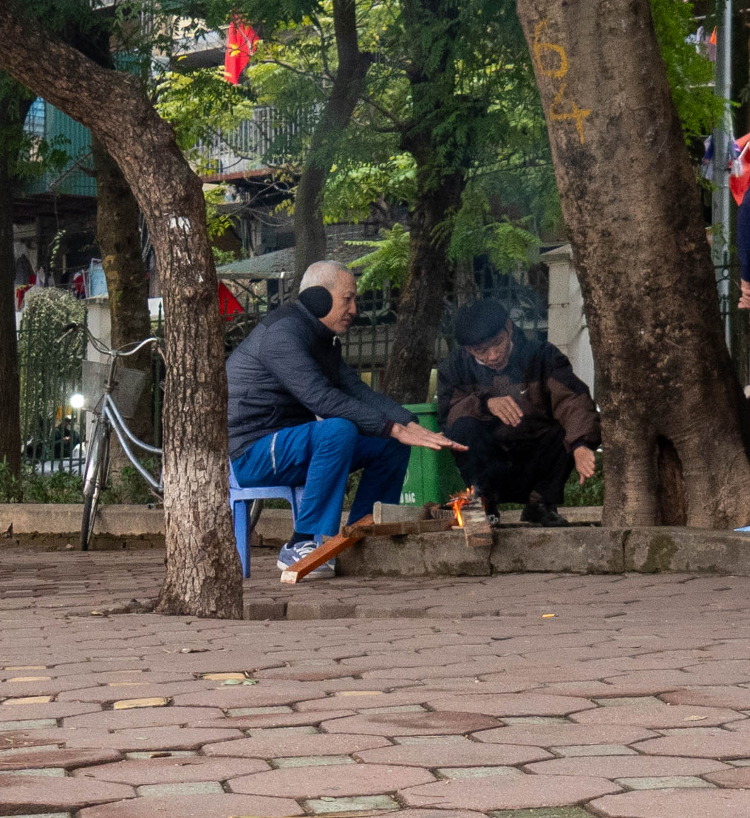 Picture of two men sitting with hands over small fire between two trees