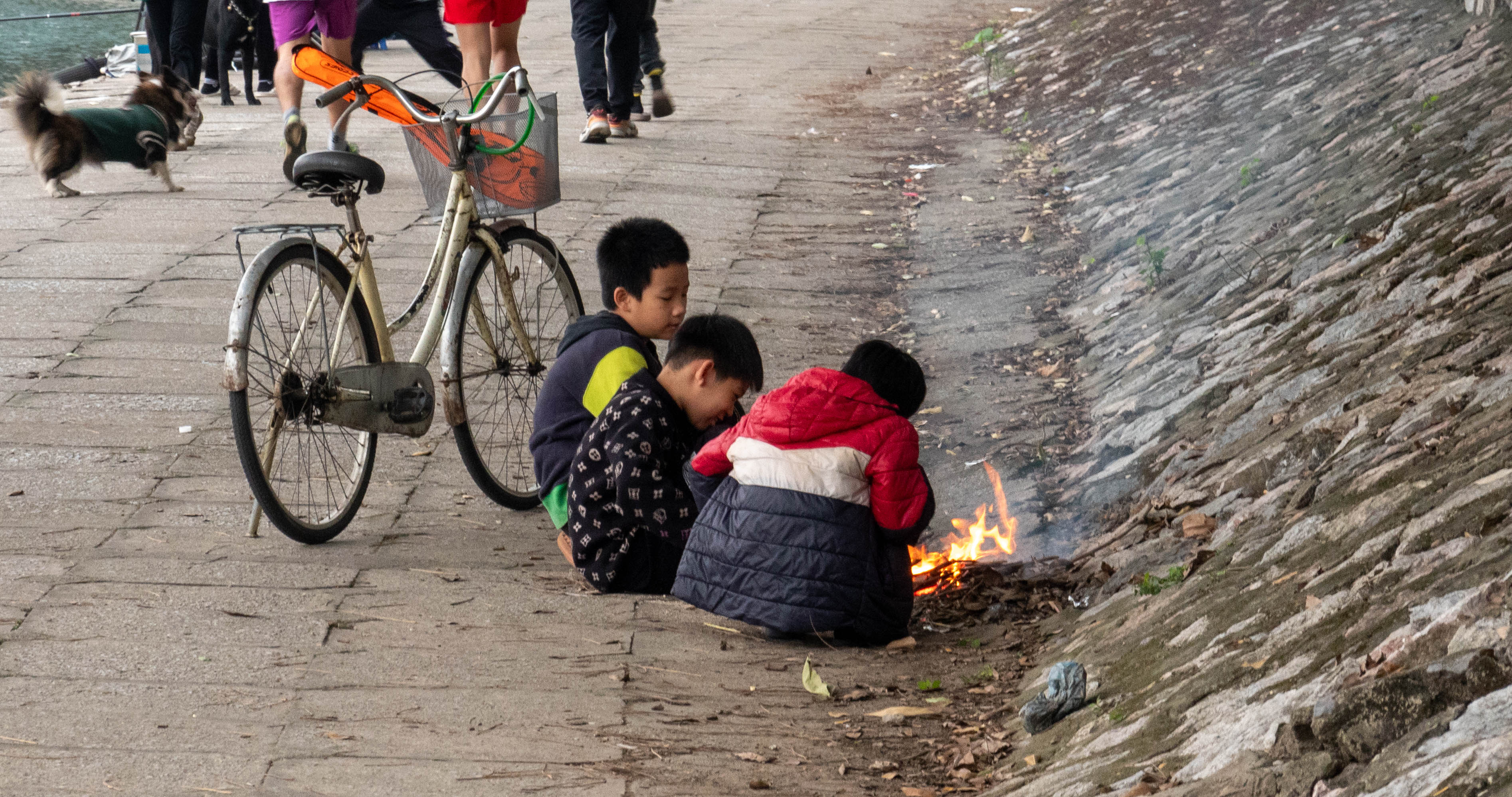 Picture of 3 you children squating in front of a small fire.  A bicycle is to the left of the picture