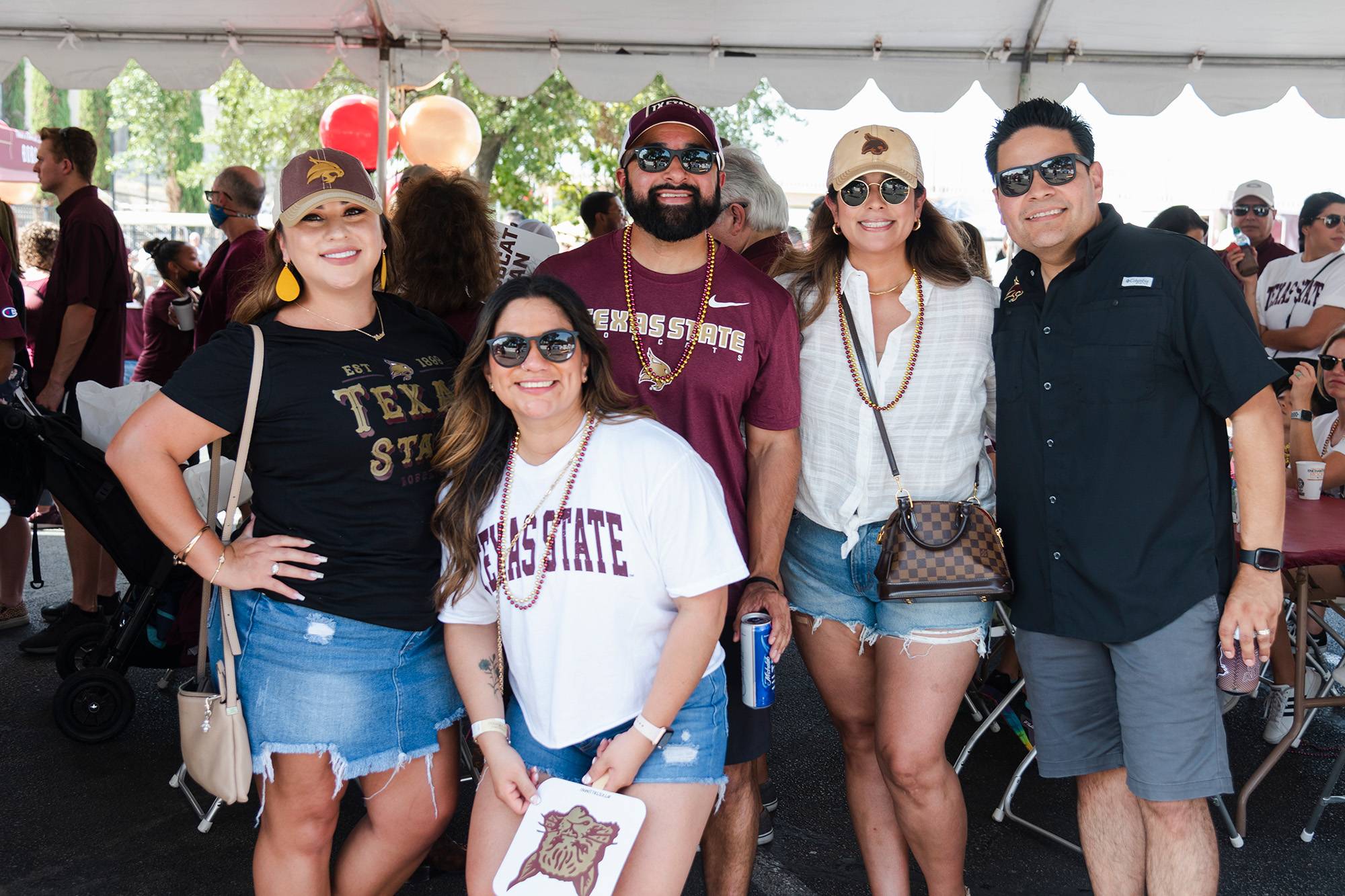 photo of five people, group smiling at TXST alumni tailgate event, wearing TXST merch