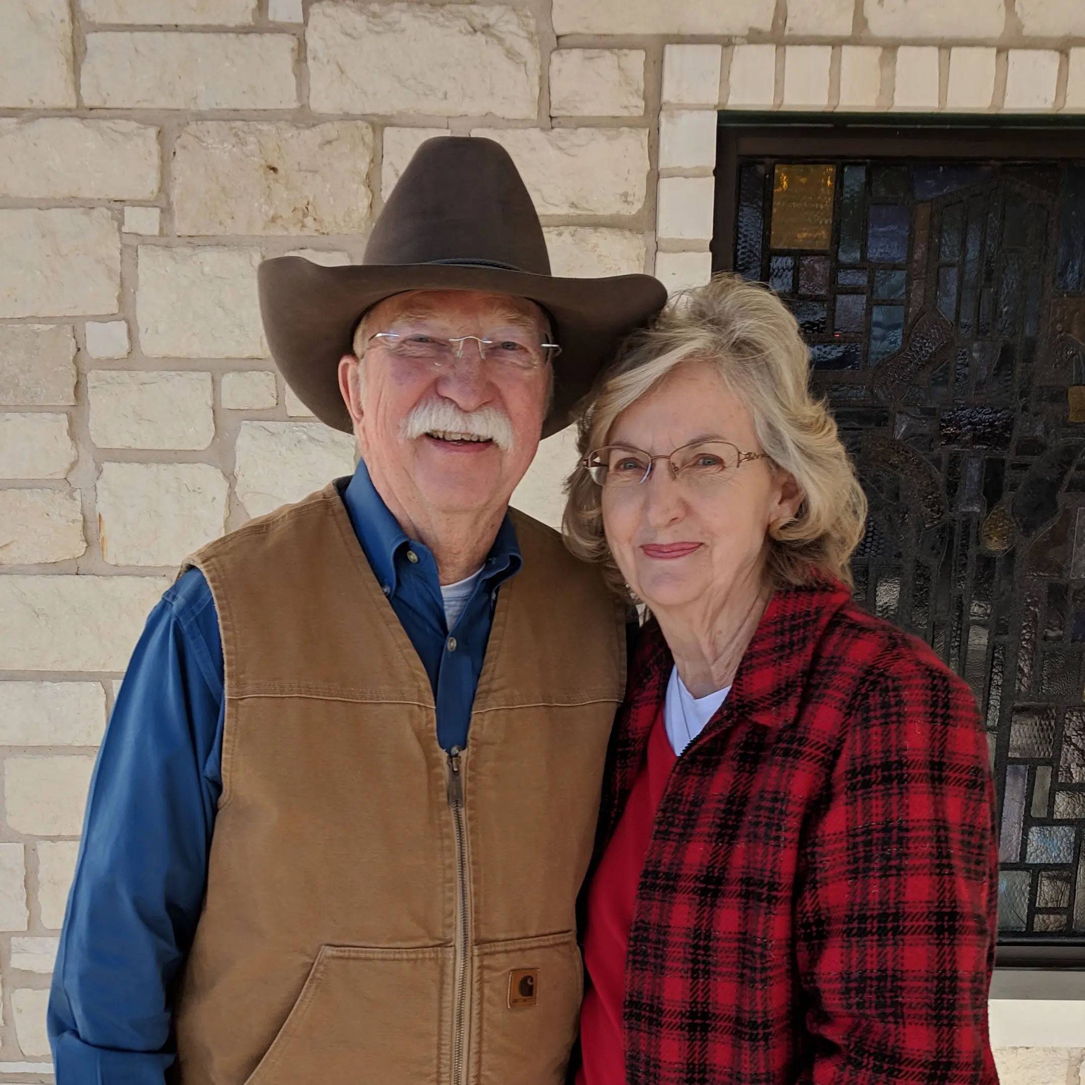 Photo of couple standing together smiling, man is wearing blue shirt and cowboy hat, woman is wearing red flannel.