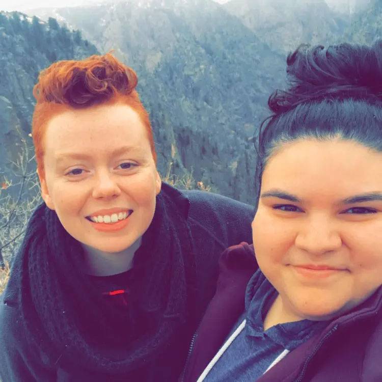 Photo of two women smiling and sitting on a cliff with trees