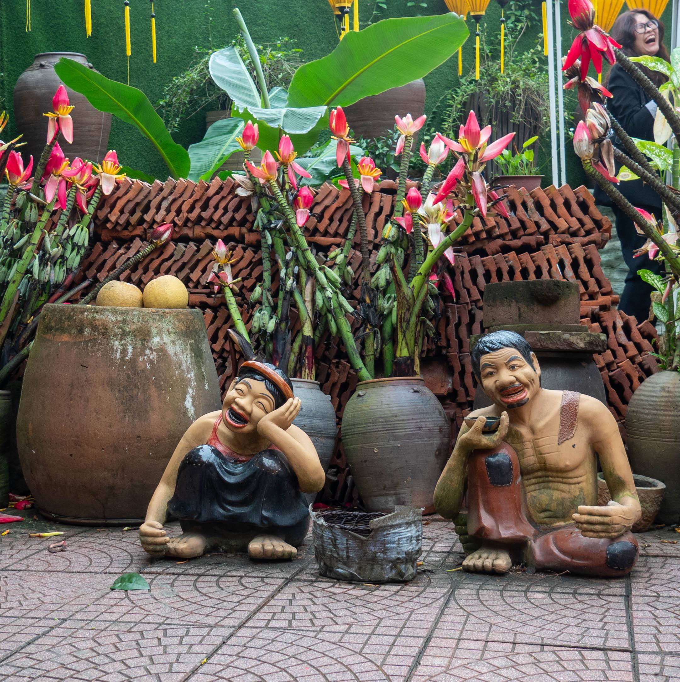 two cartoon-like men sitting with a gray urn between them flowers in the background 