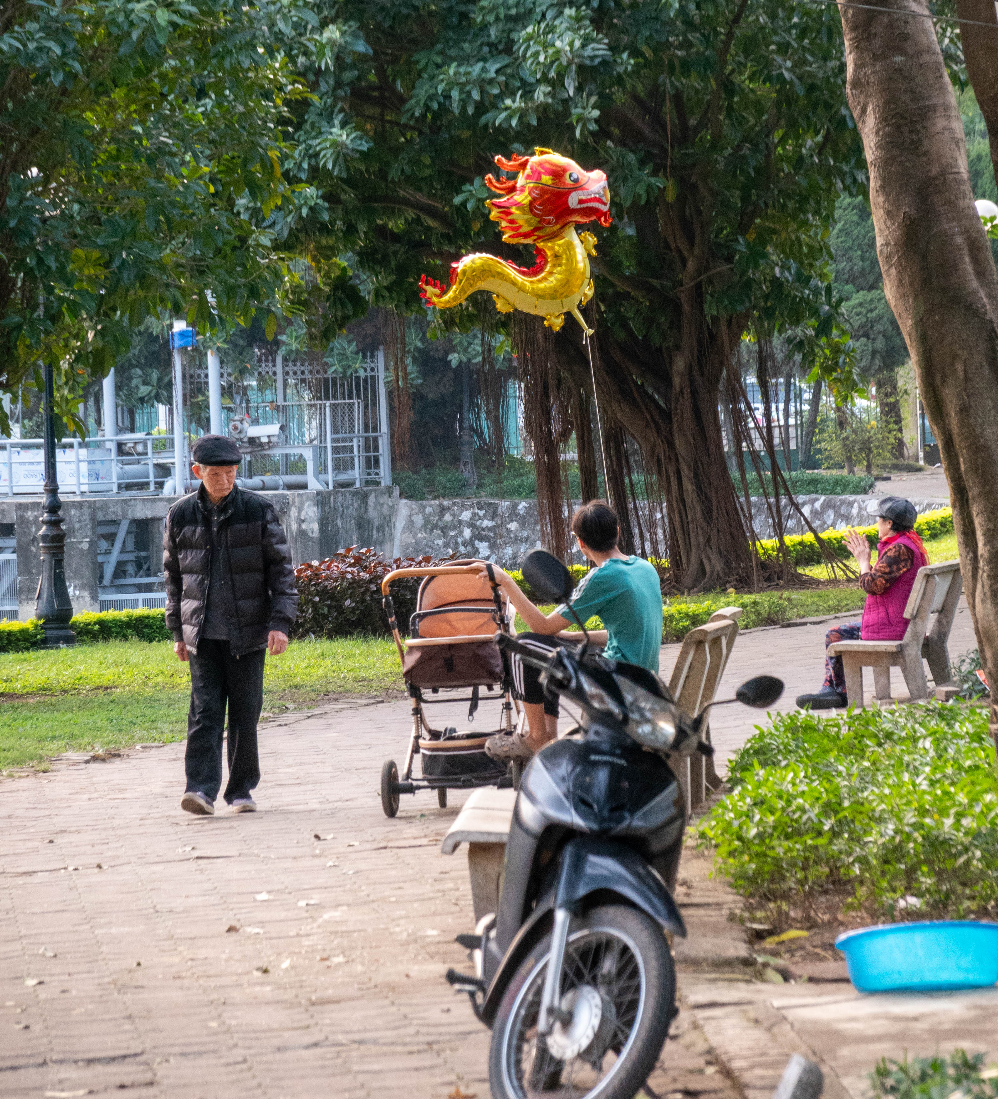 Picture of elderly man (on left) left walking by a man sitting on a bench with his feet propped up on a baby carrigage holding a mylar helium filled dragon who is looking at an elderly lady sitting on a bench on the right of picture