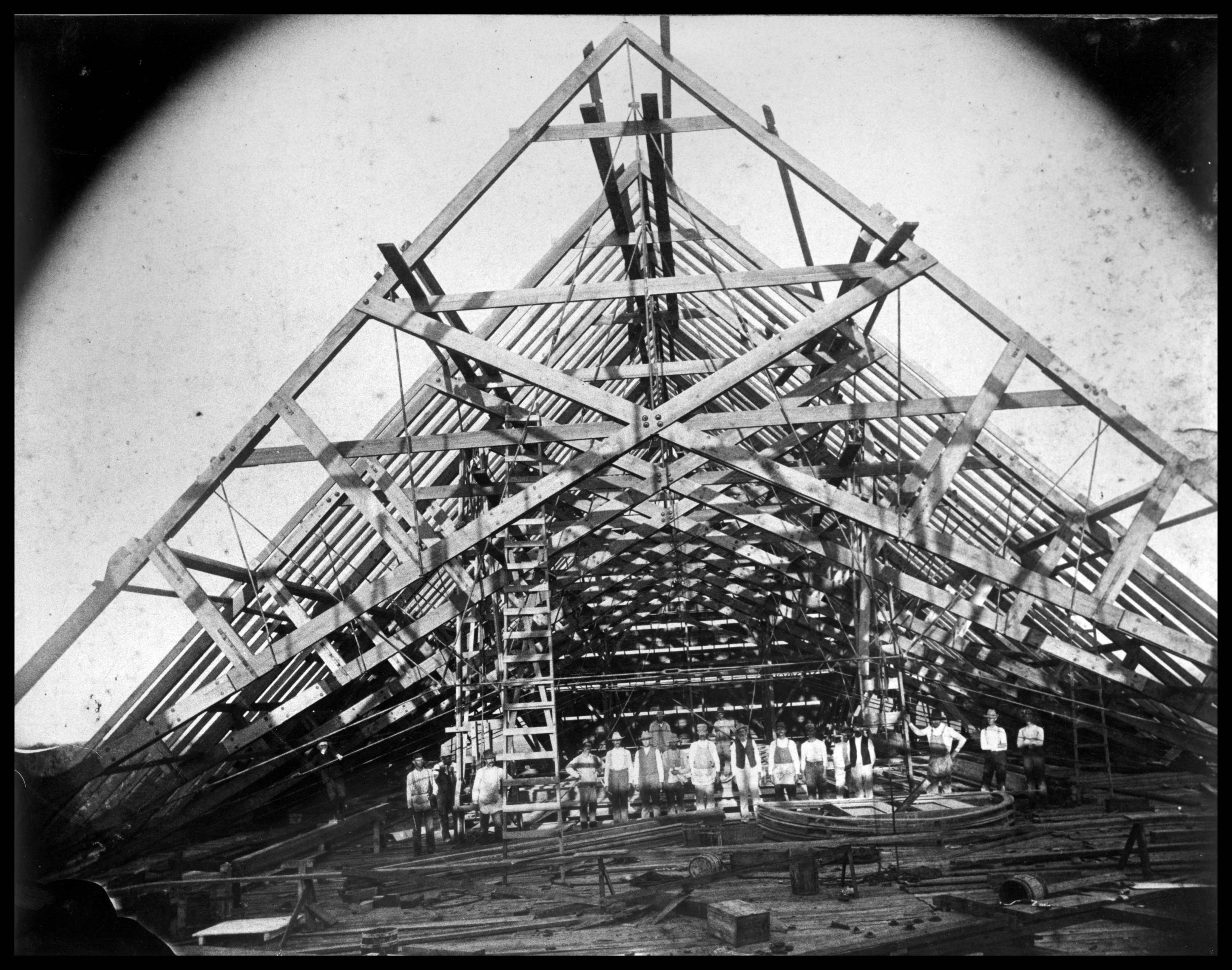 Aged black and white photograph of Old Main's roof under construction. A crew of men are lined up to be photographed under the open roof beams.